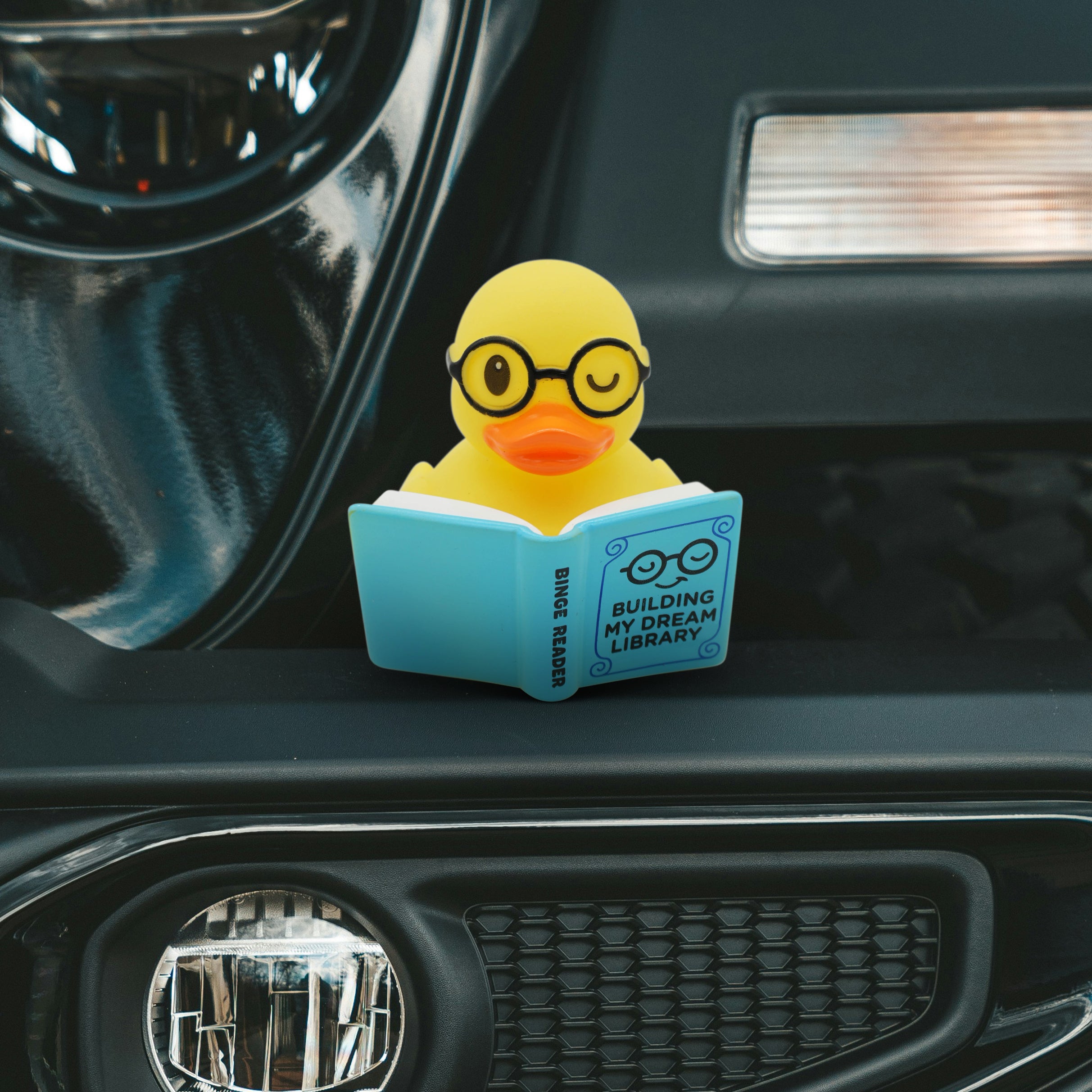 Yellow rubber duck with glasses reading a book on a car dashboard