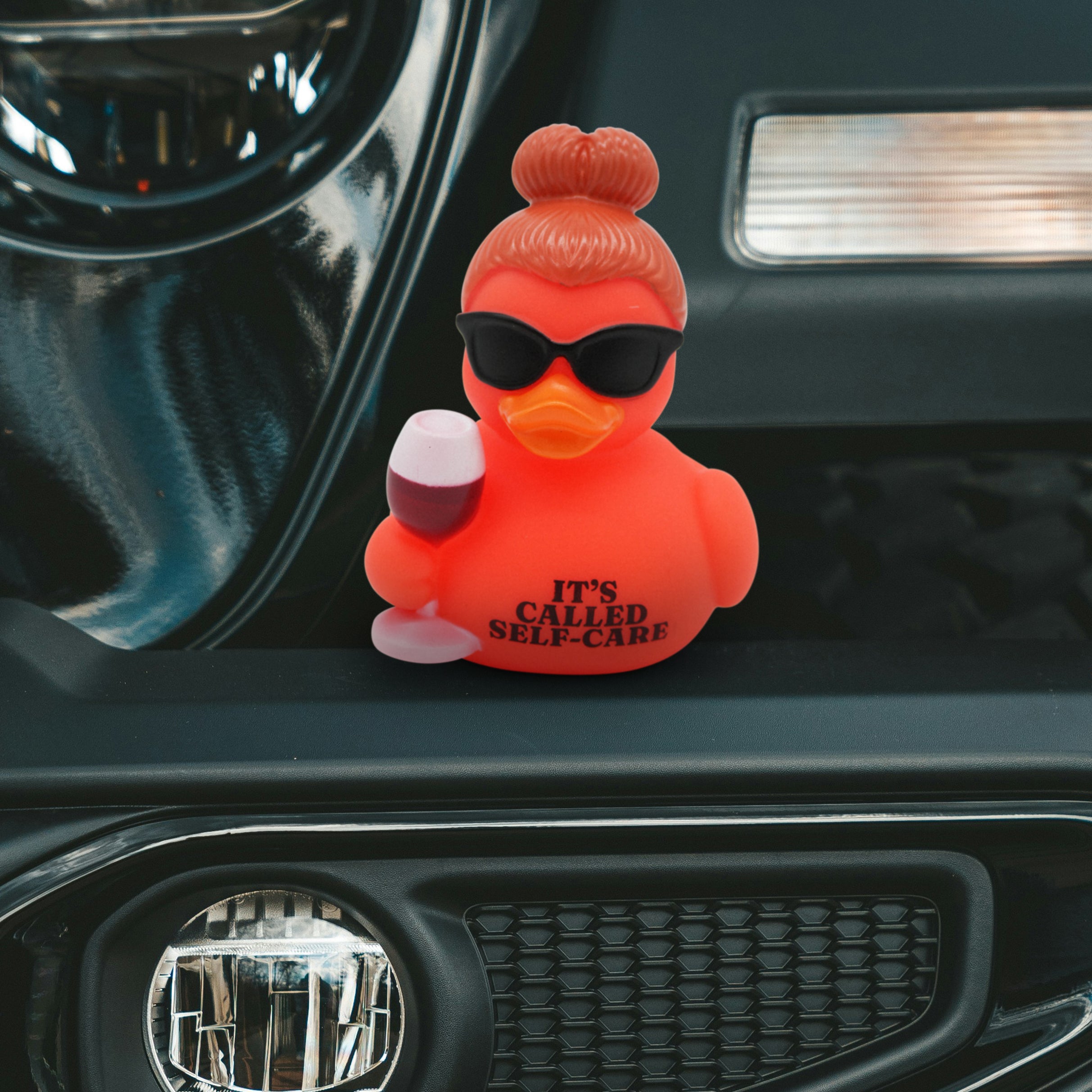 Red rubber duck with sunglasses and a glass of wine with the text 'It's Called Self-Care' on a car dashboard.