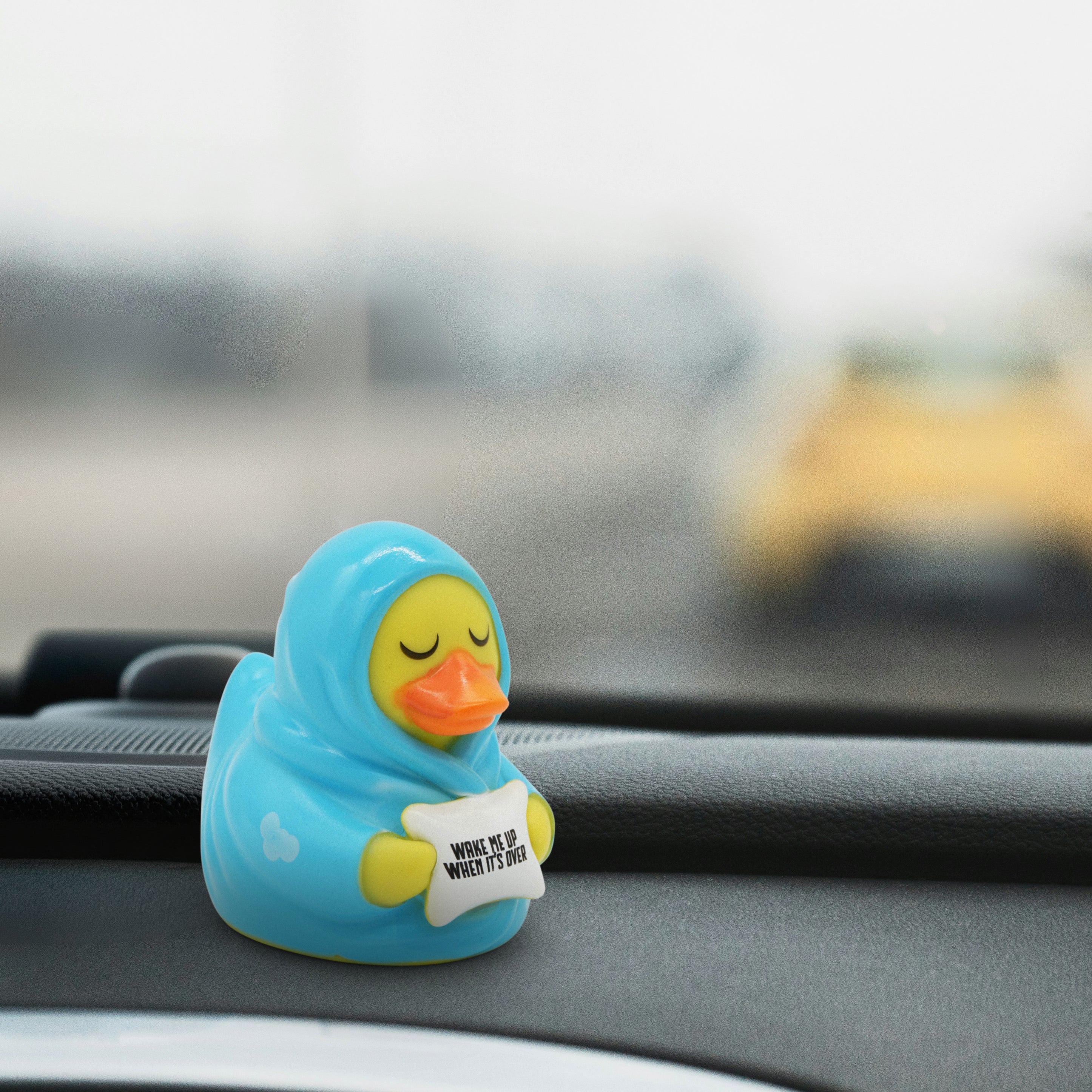 Rubber duck toy in a blanket holding a pillow that reads "Wake me up when it's over" on a car dashboard.