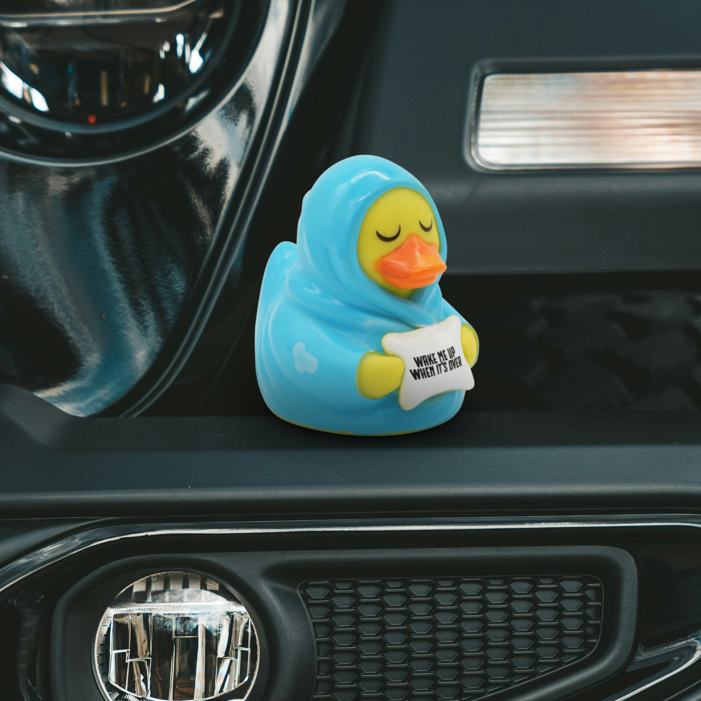 Rubber duck toy in a blanket holding a pillow that reads "Wake me up when it's over" on a car dashboard.