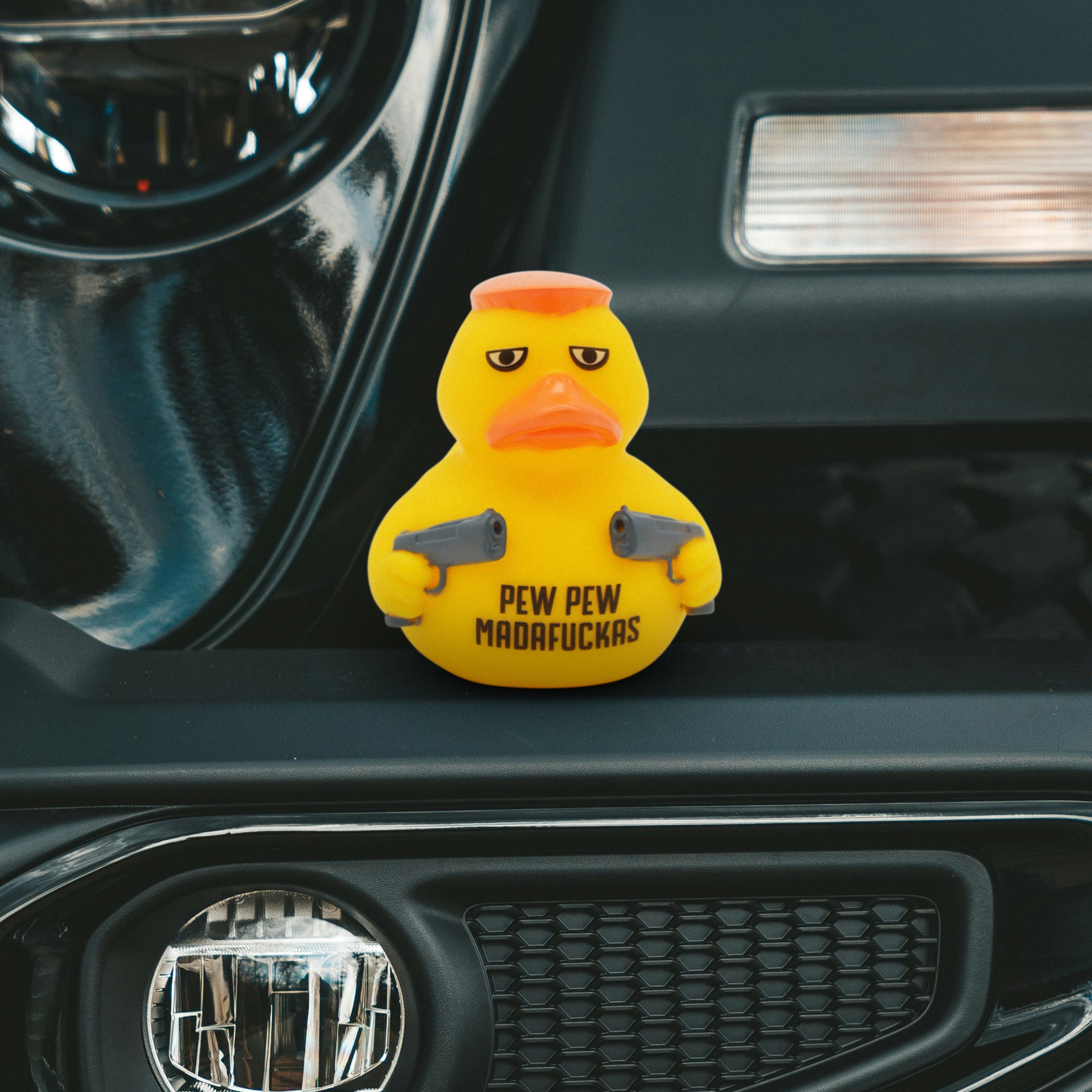 Yellow rubber duck with a serious expression holding guns with the text "pew pew madafucka" written on it on a car dashboard.
