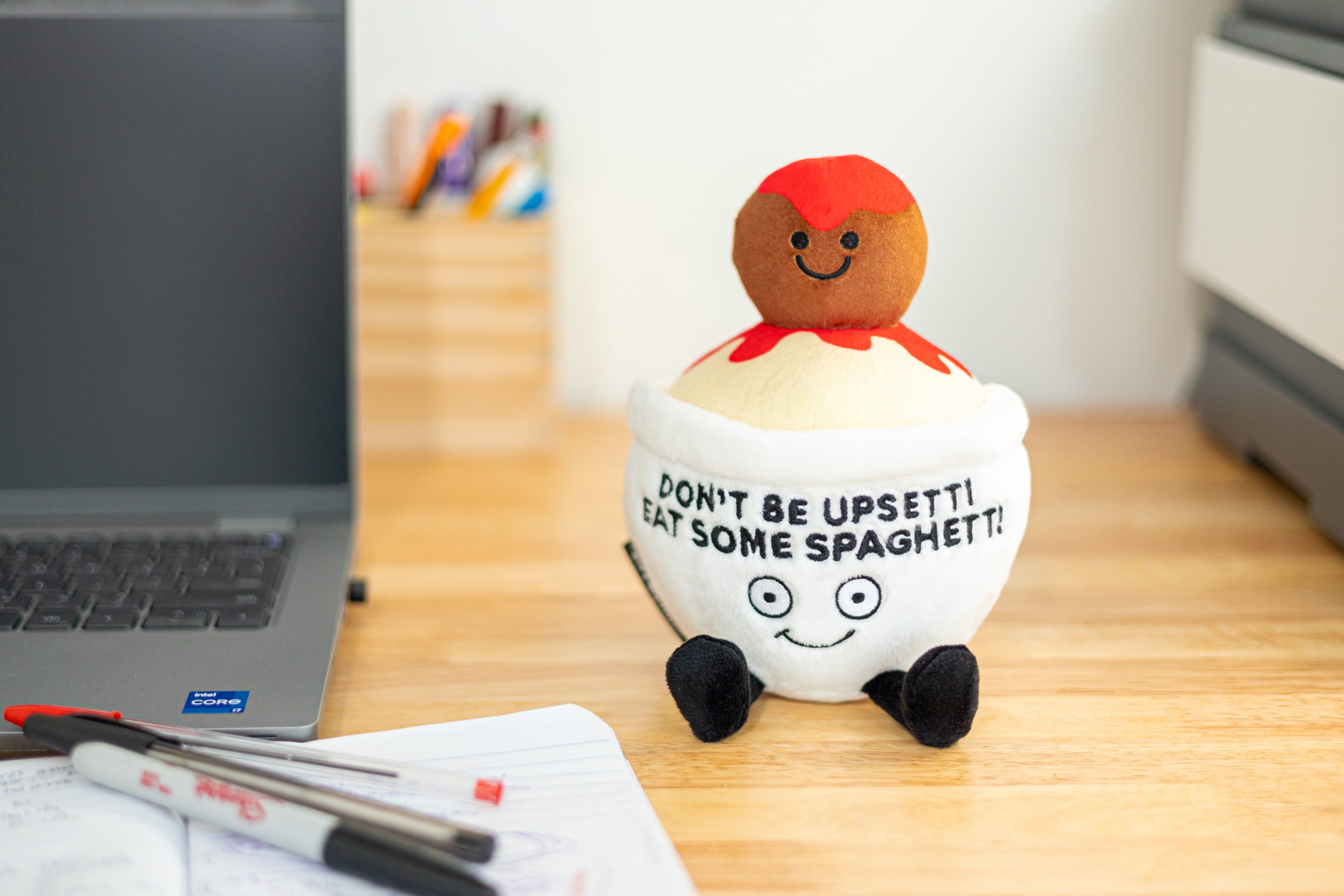 A plush resembling a bowl of spaghetti with a smiling meatball on top that reads, 'DON'T BE UPSETTI EAT SOME SPAGHETTI'. It's placed on a desk near a computer.