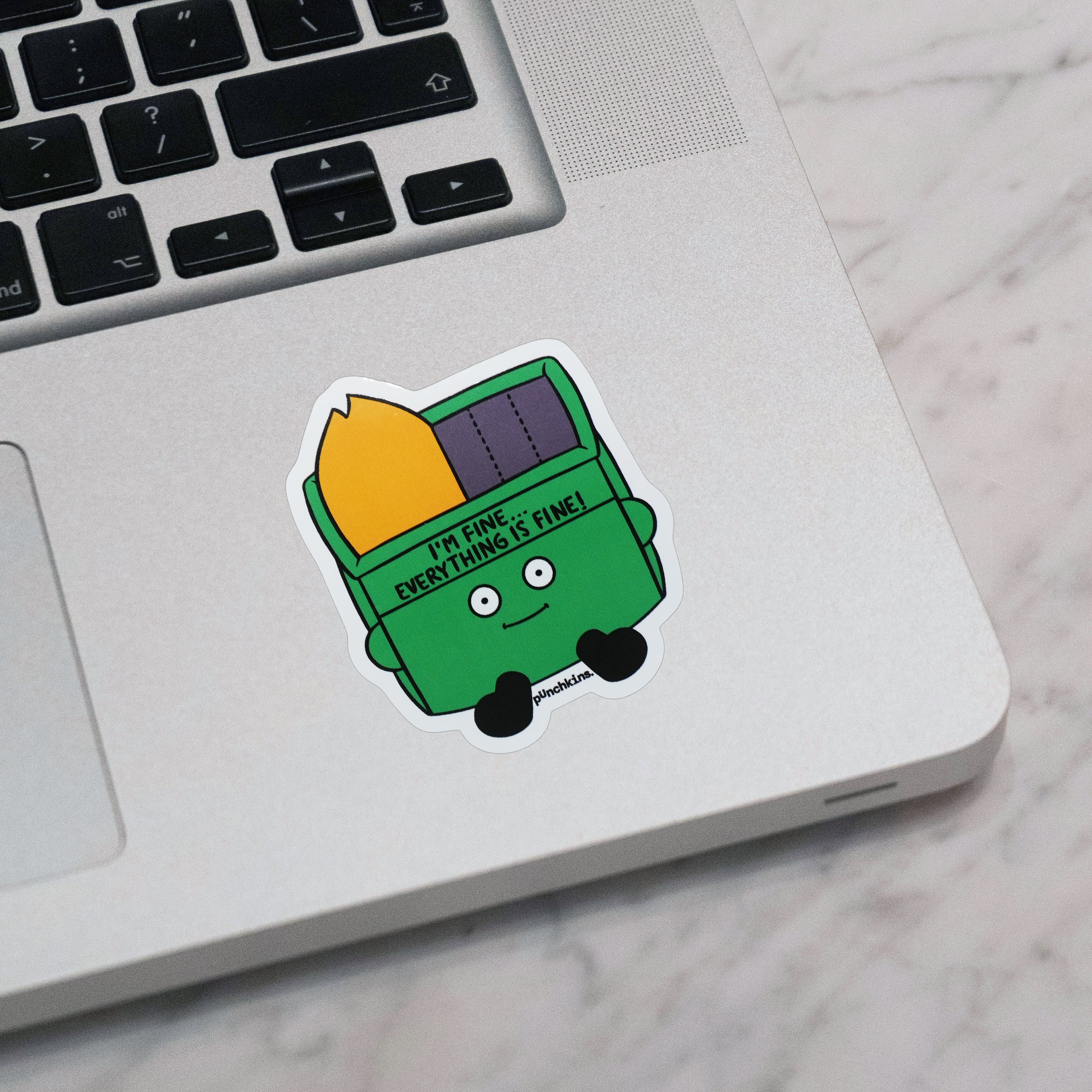 A die-cut sticker of a green cartoon dumpster that's on fire placed on the corner of a silver laptop on a white marble surface. The sticker features the text 'I'M FINE...EVERYTHING IS FINE!'