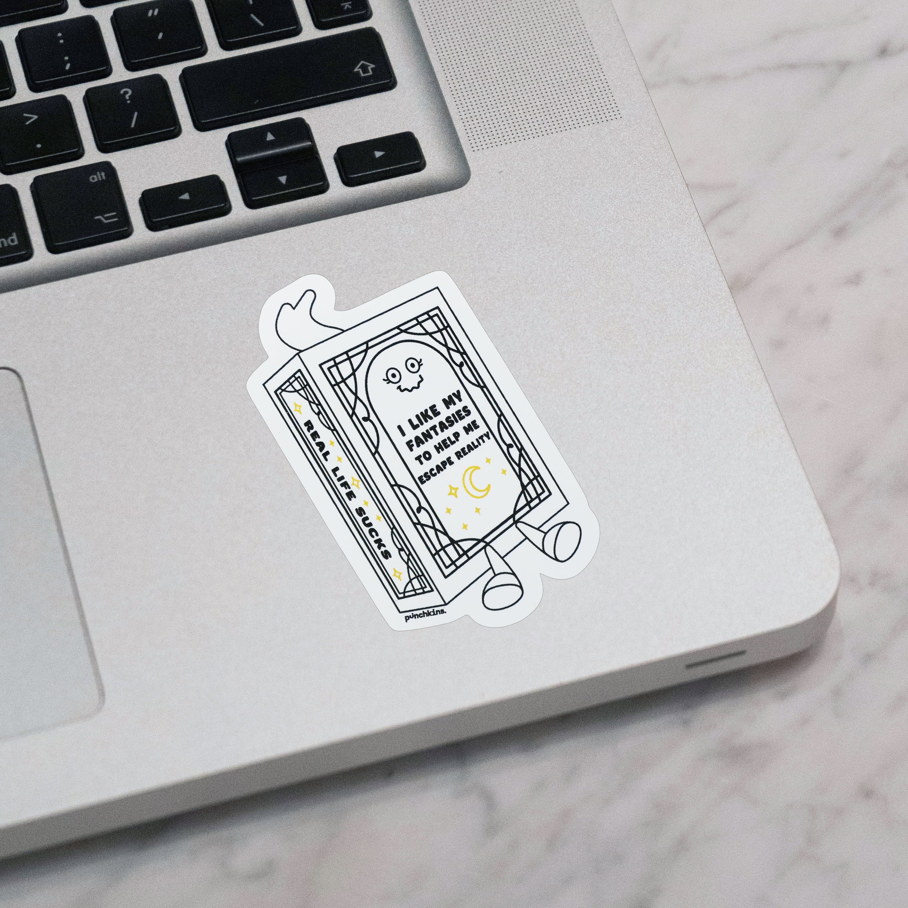 A die-cut vinyl sticker featuring a cartoon book character with the text 'I LIKE MY FANTASY TO HELP ME ESCAPE REALITY' applied to the corner of a silver laptop, set on a light gray marble surface.