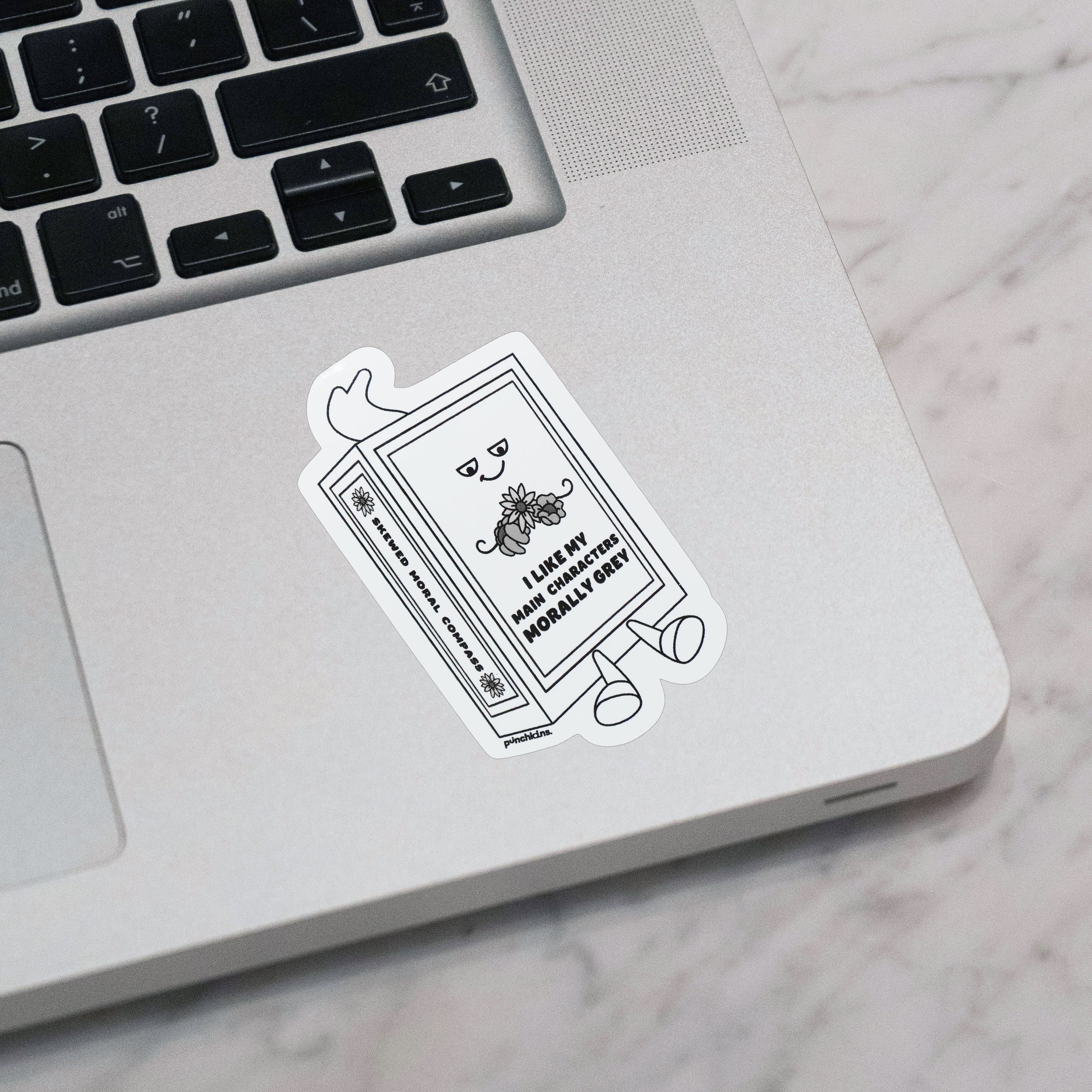 A die-cut vinyl sticker featuring a cartoon book character with the text 'I LOVE MY MAIN CHARACTERS MORALLY GREY' applied toa silver laptop on a white marble surface.