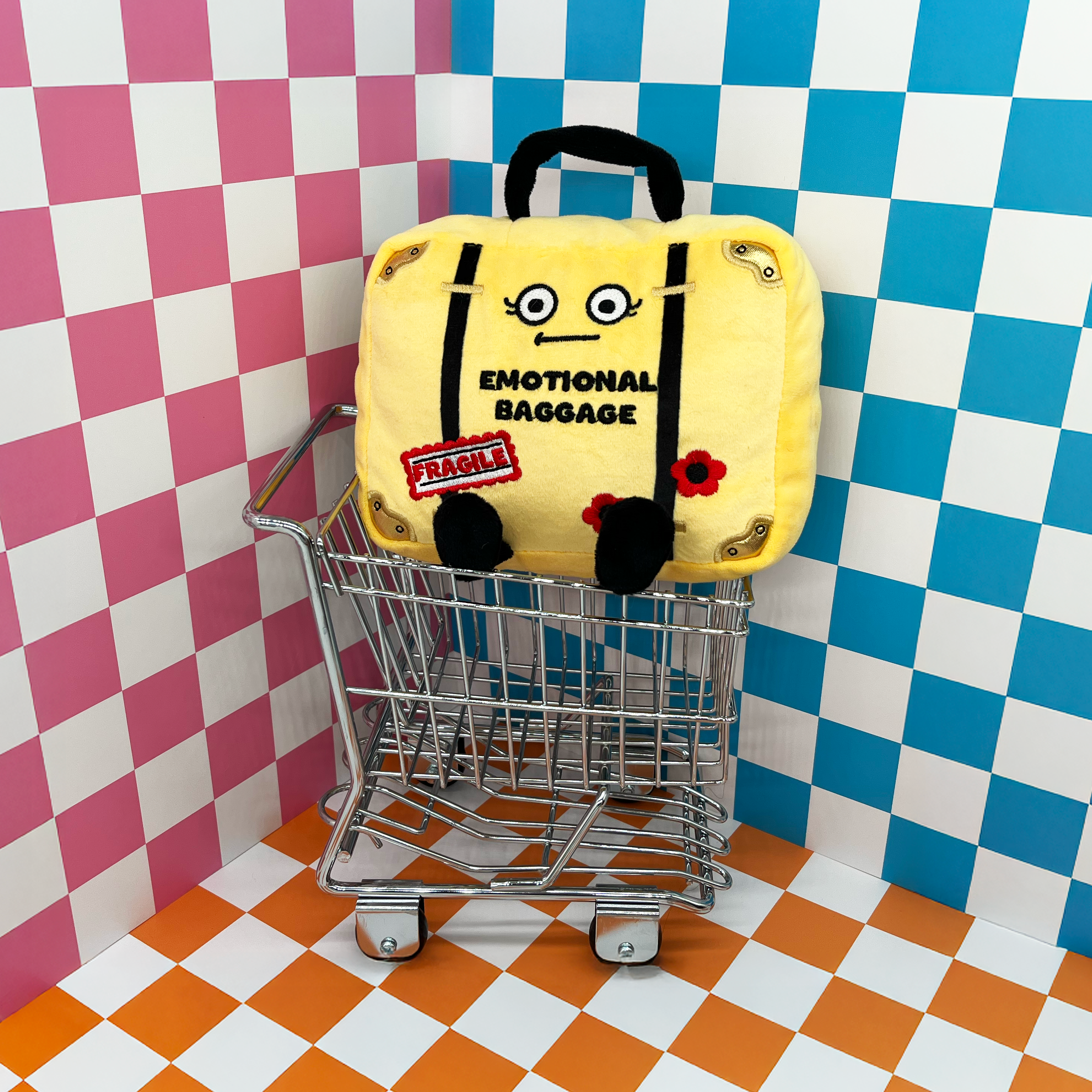 Yellow suitcase labeled 'Emotional Baggage' in a small shopping cart against a checkered wall.