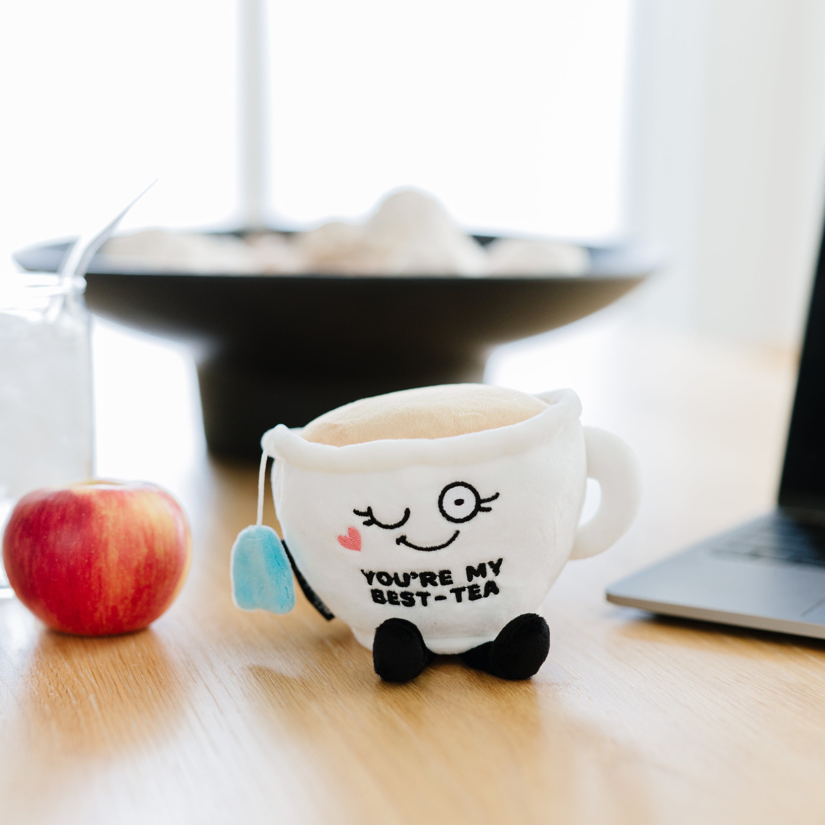 A plush resembling a white teacup featuring a smiling face and the text 'YOU'RE MY BEST-TEA' written on it placed on a kitchen table.