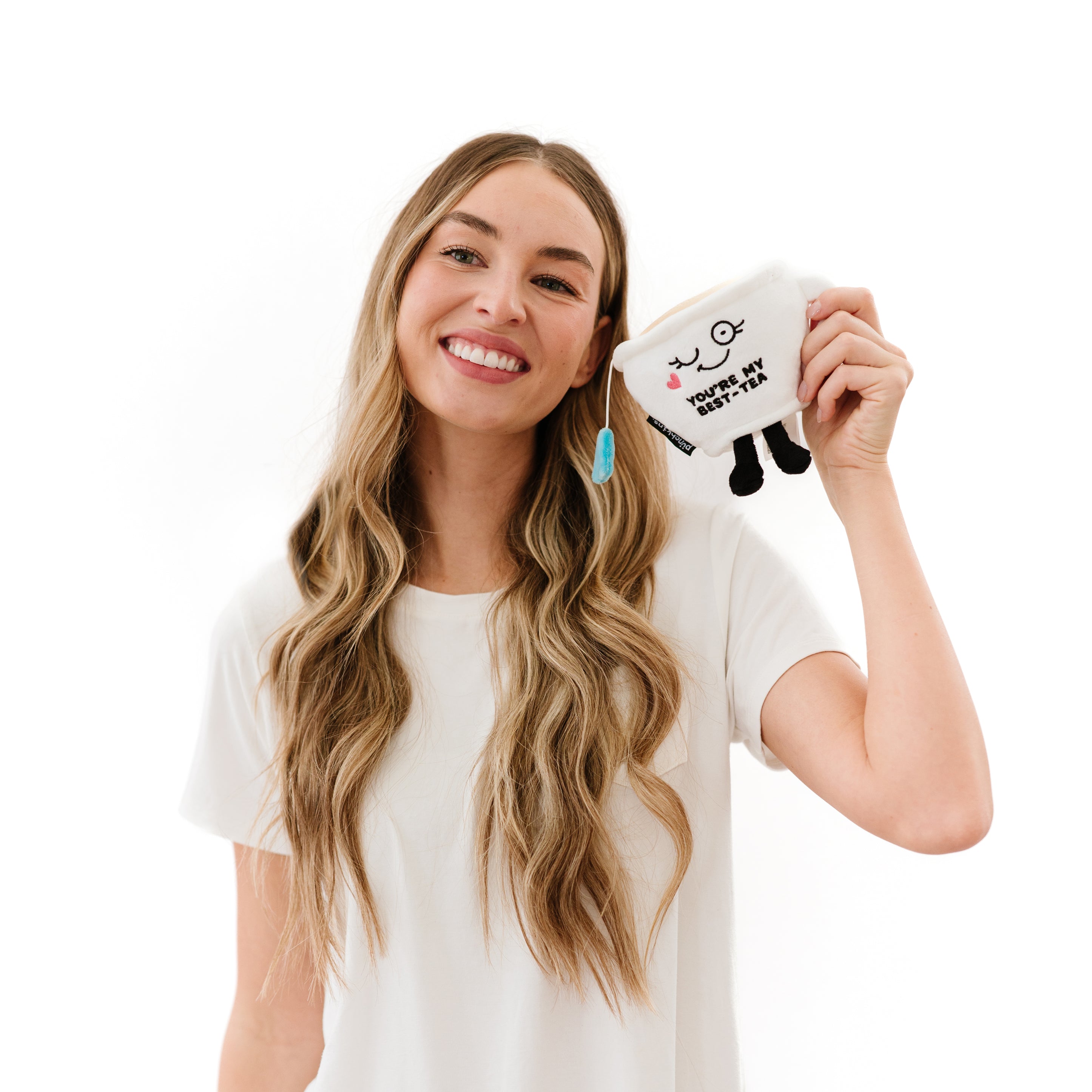 A woman holding a plush resembling a white teacup featuring a smiling face and the text 'YOU'RE MY BEST-TEA' written on it.