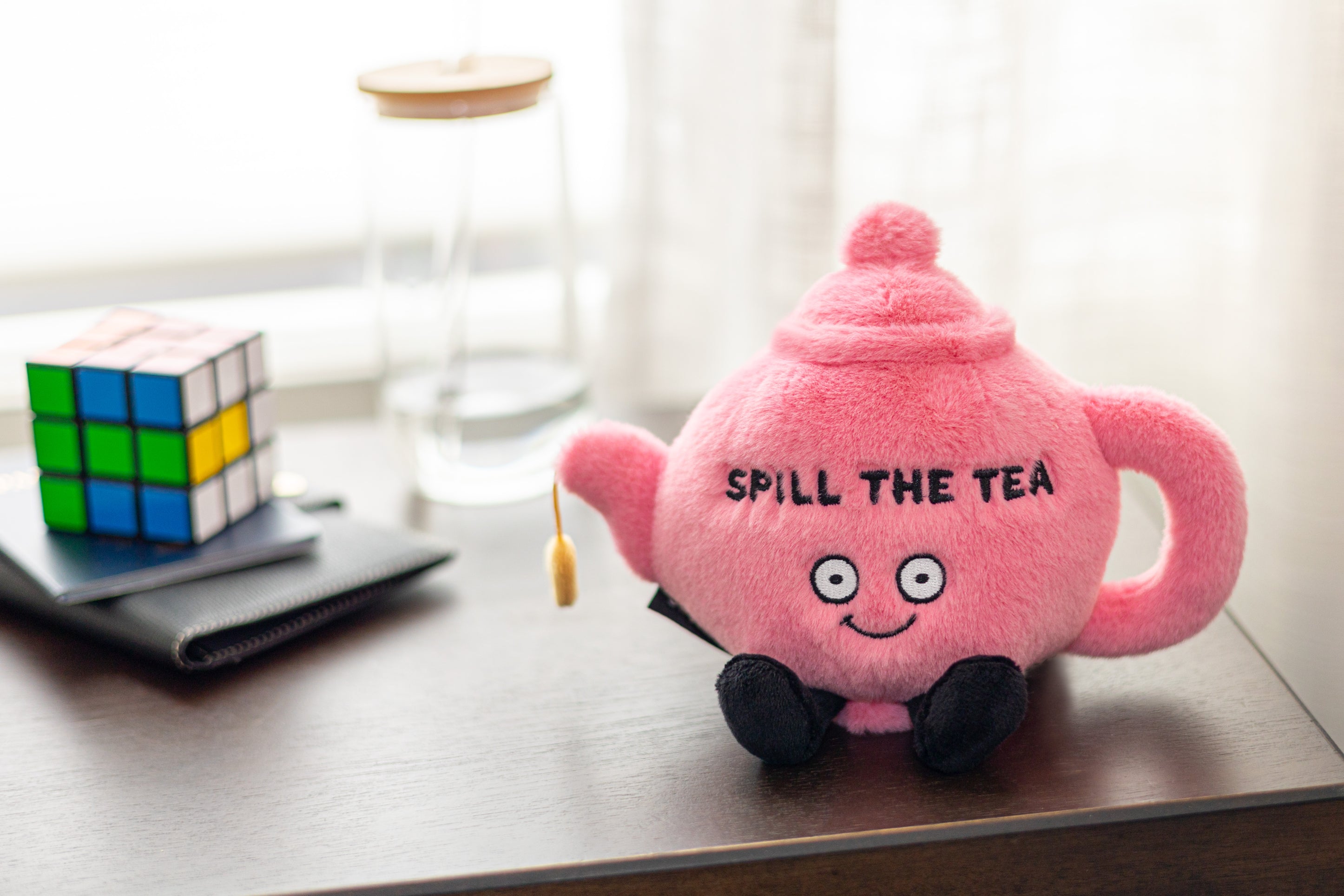 A plush pink teapot with the phrase 'SPILL THE TEA' written on it sitting on a shelf.