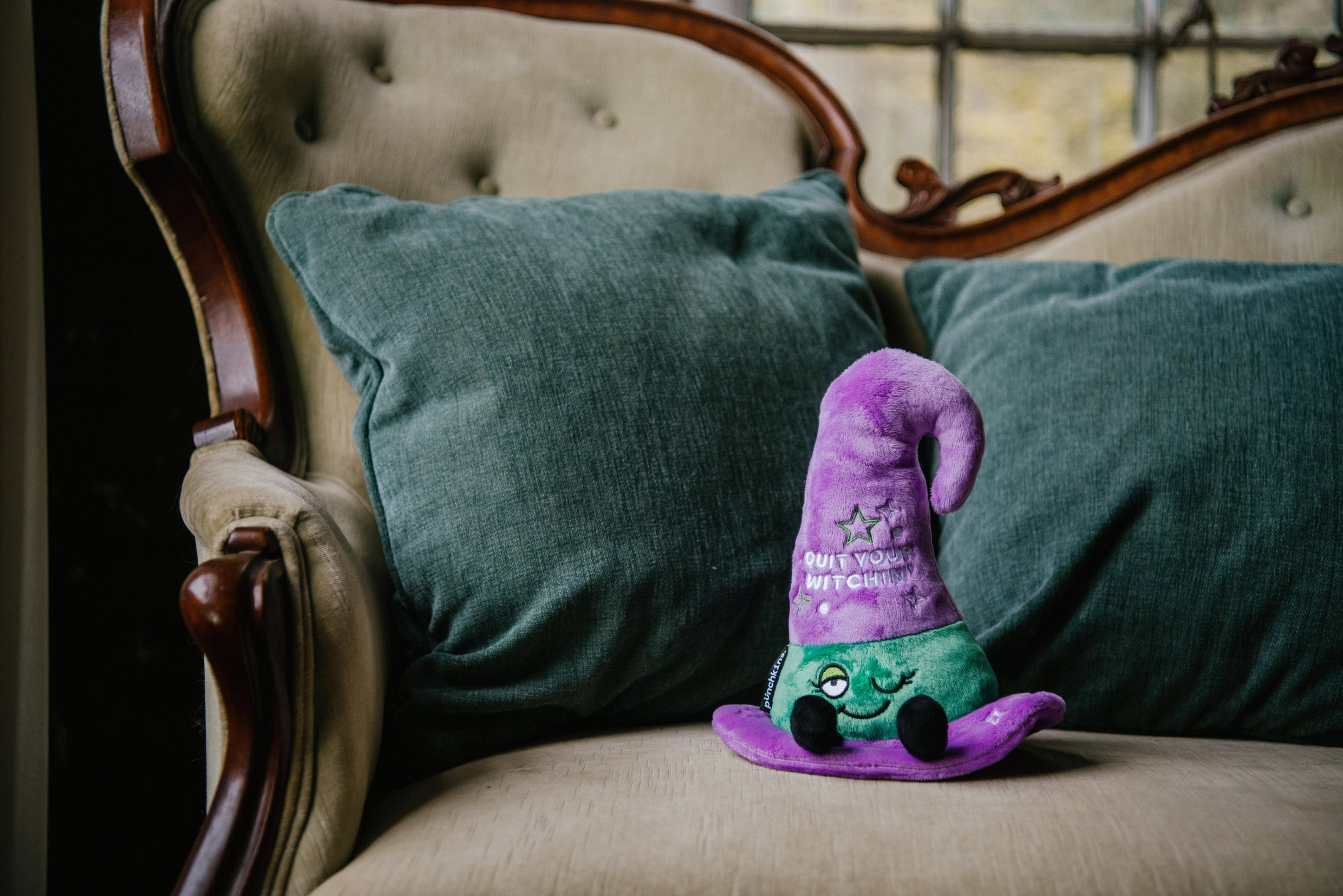 A plush witch hat featuring text that reads, "quit your witching'" placed on a couch.