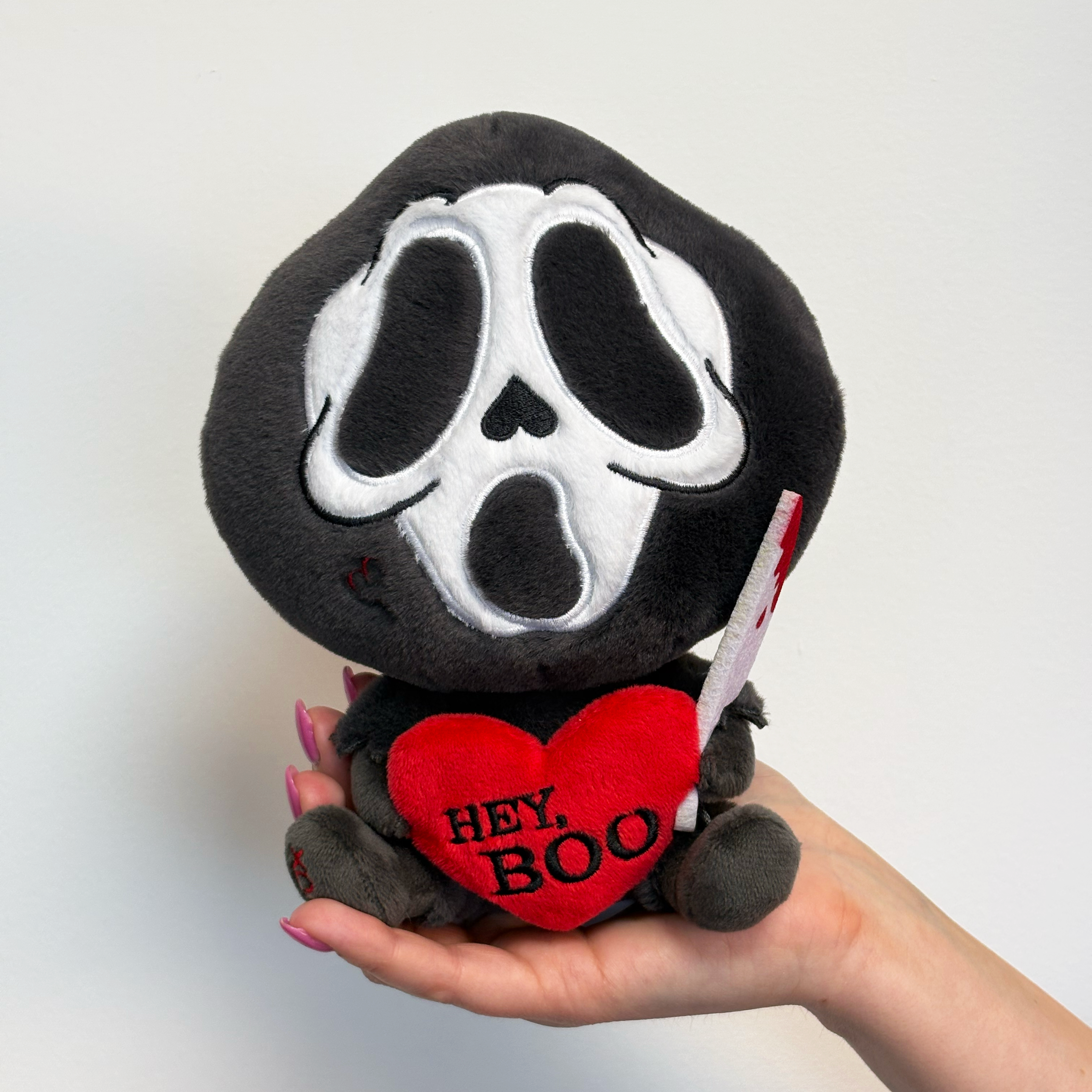 A hand holding ghost face killer plush that says, "Hey, boo," in a heart.