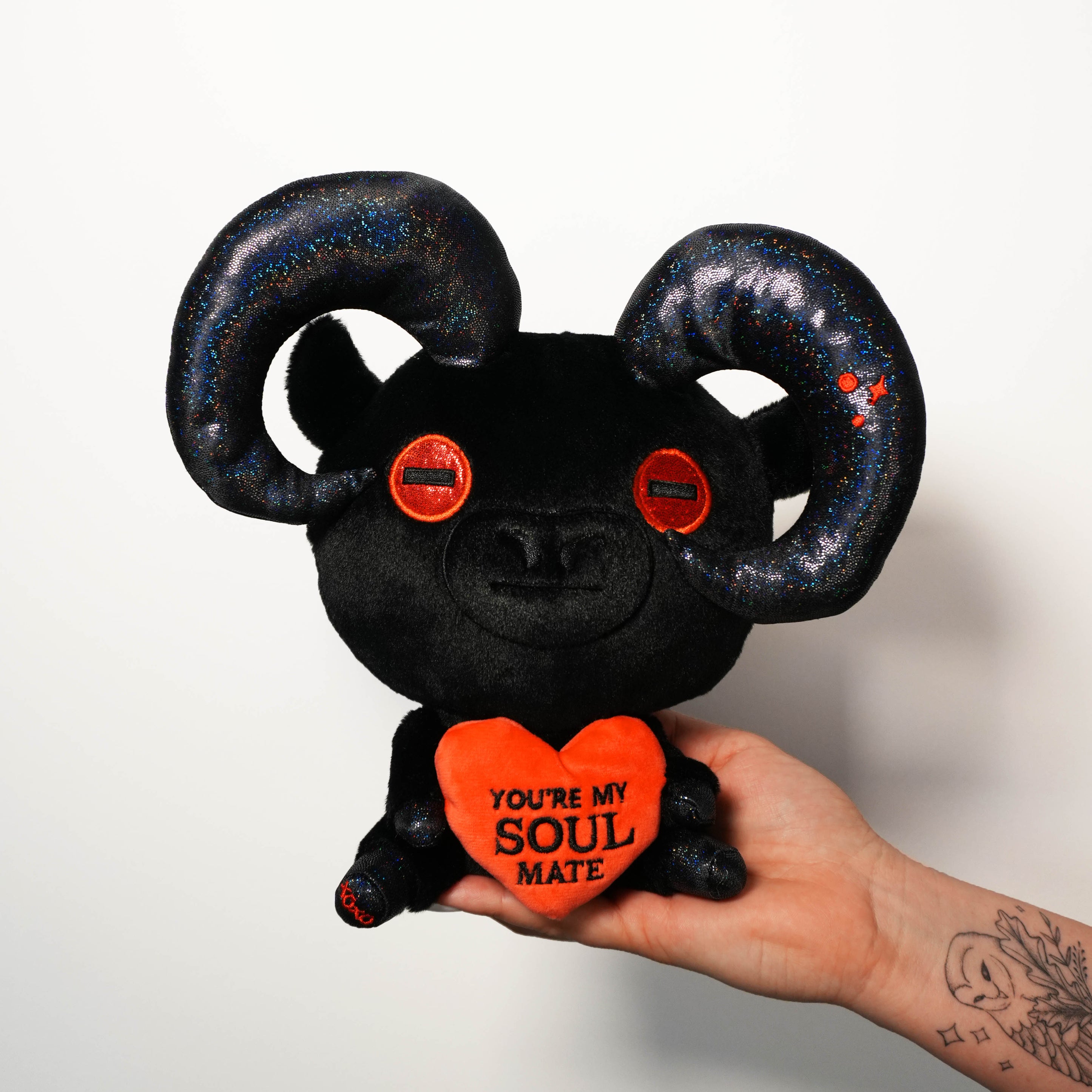 Devil goat plush toy with red heart and text held by a hand against a white background