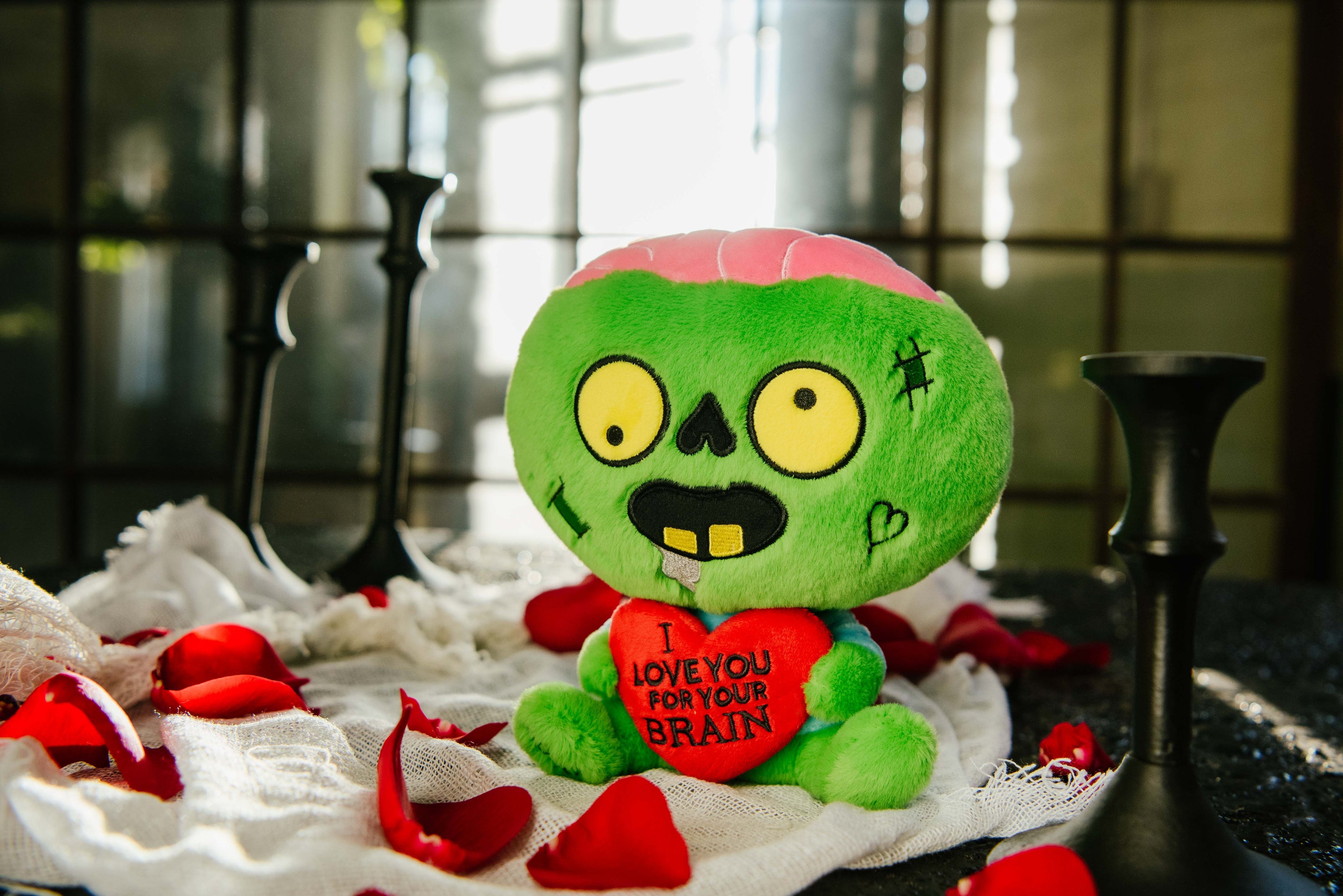 A zombie plush with a 3D heart that reads, "I love you for your brains," sitting on a bed of rose petals.