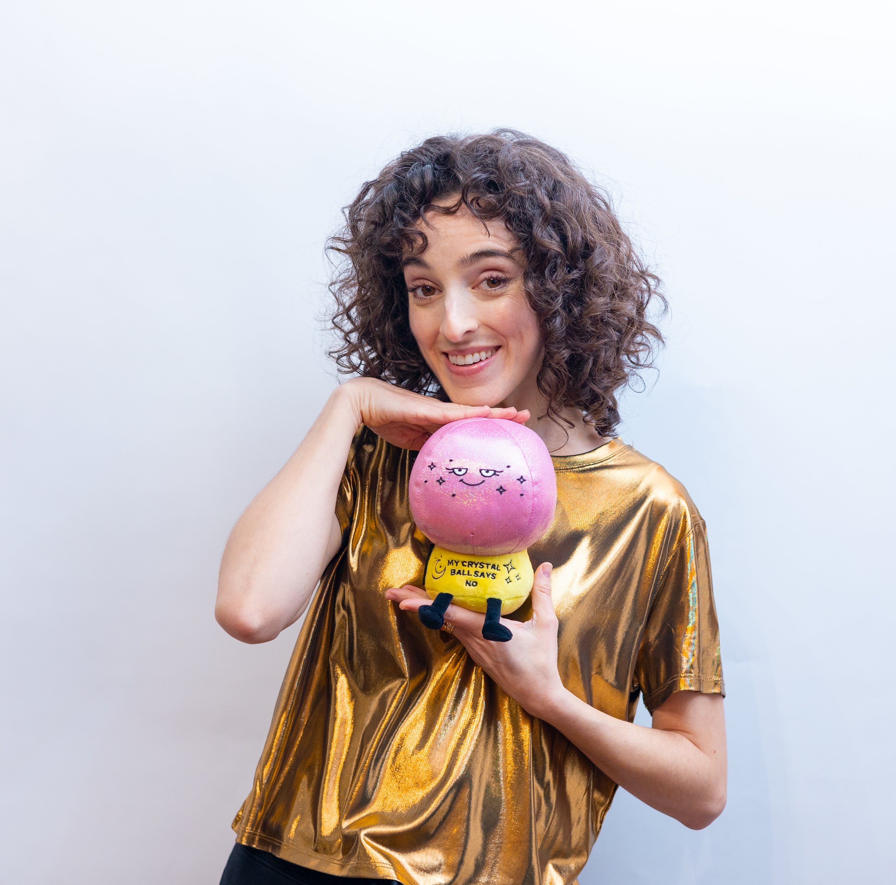 A woman is holding a plush resembling a crystal ball with a pink sparkly top and a cute face and has text that reads 'MY CRYSTAL BALL SAYS NO'.