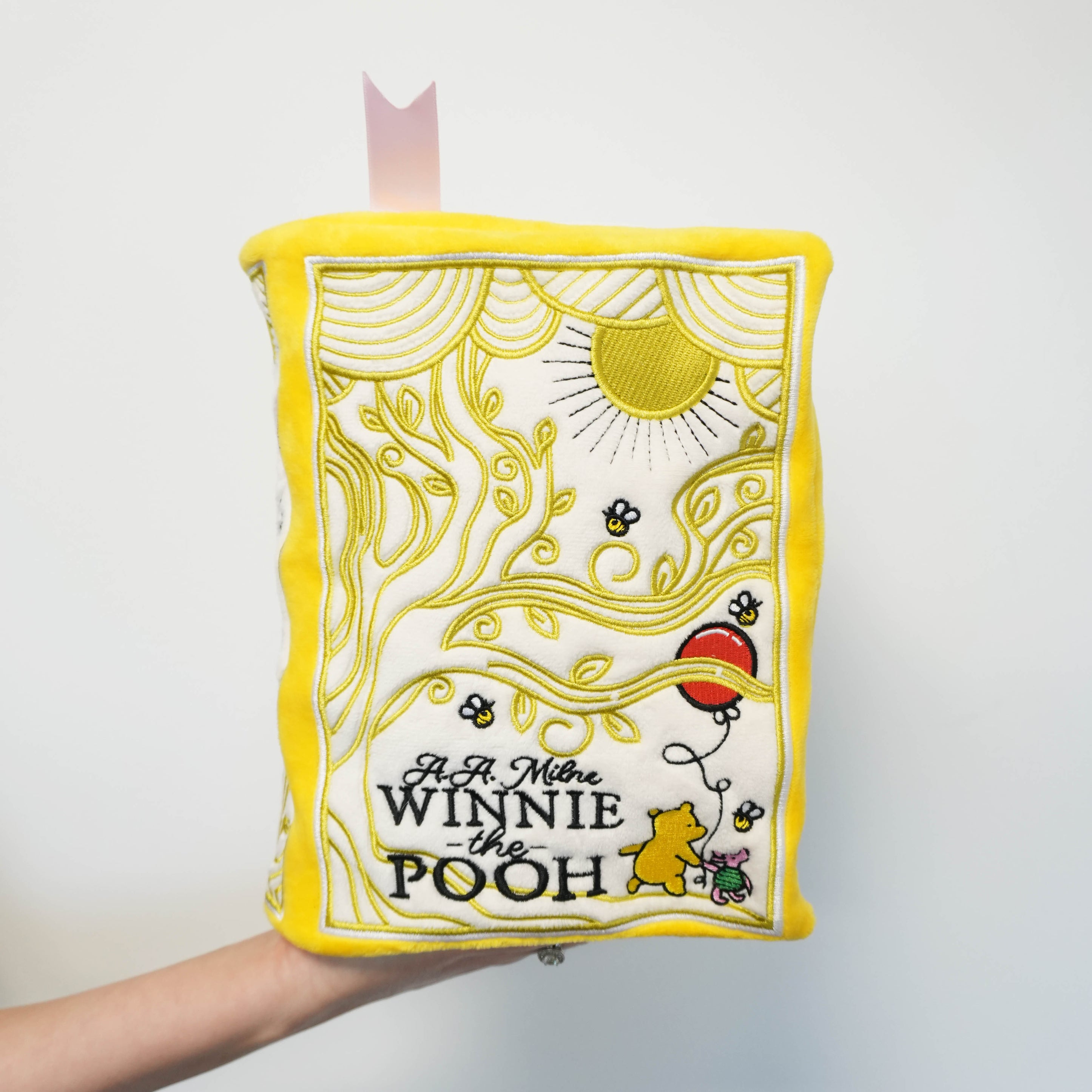Hand holding a book-shaped plush with 'Winnie the Pooh' design on a white background