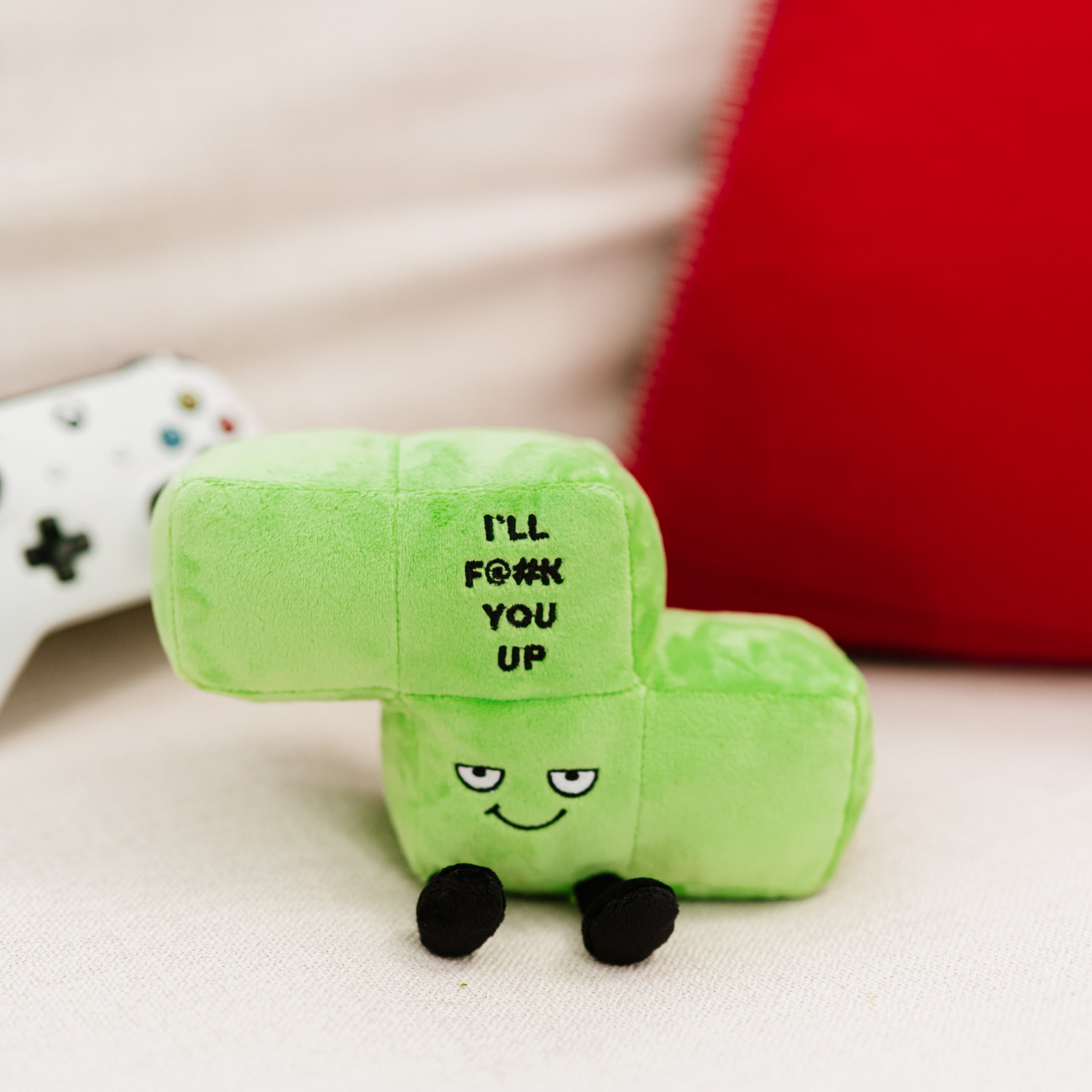 A plush designed to look like a green block with a mischievous expression and the phrase 'I'll F@#K you up' written on it placed on a couch.