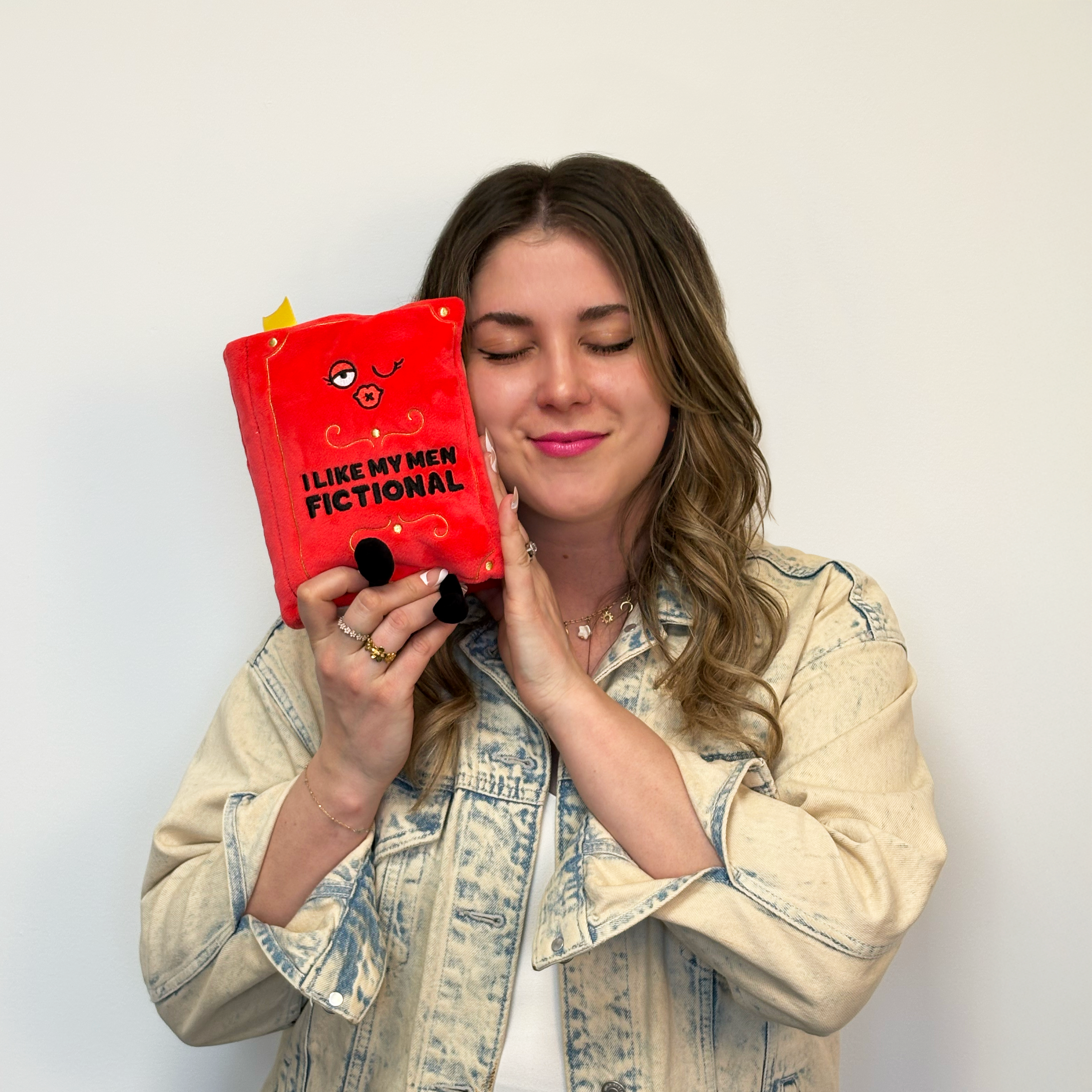 A woman holding a plush designed to look like a red book with 'I LIKE MY MEN FICTIONAL' printed on the cover.