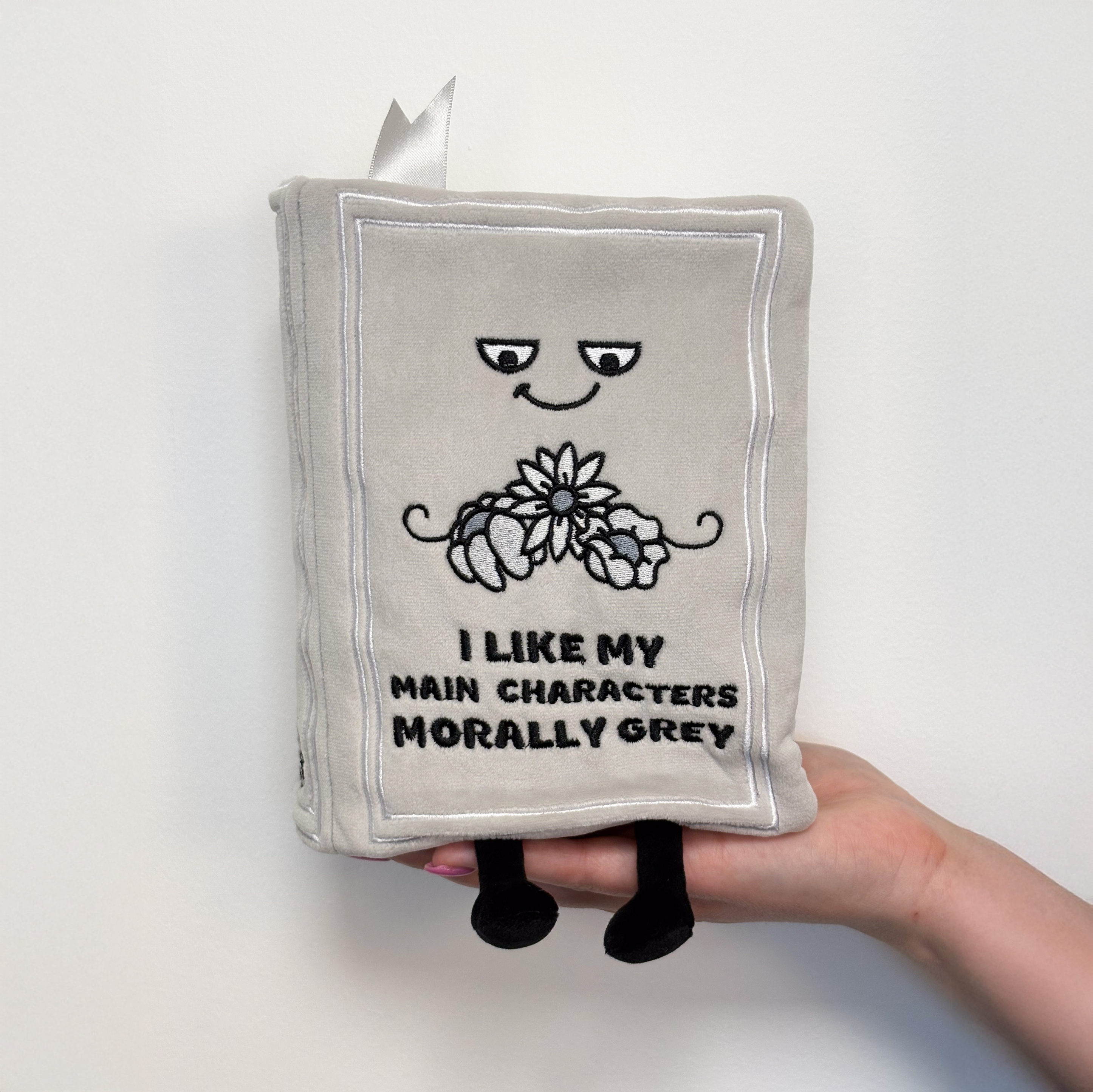 Hand holding a gray plush book with the text 'I LIKE MY MAIN CHARACTERS MORALLY GREY' on the cover.