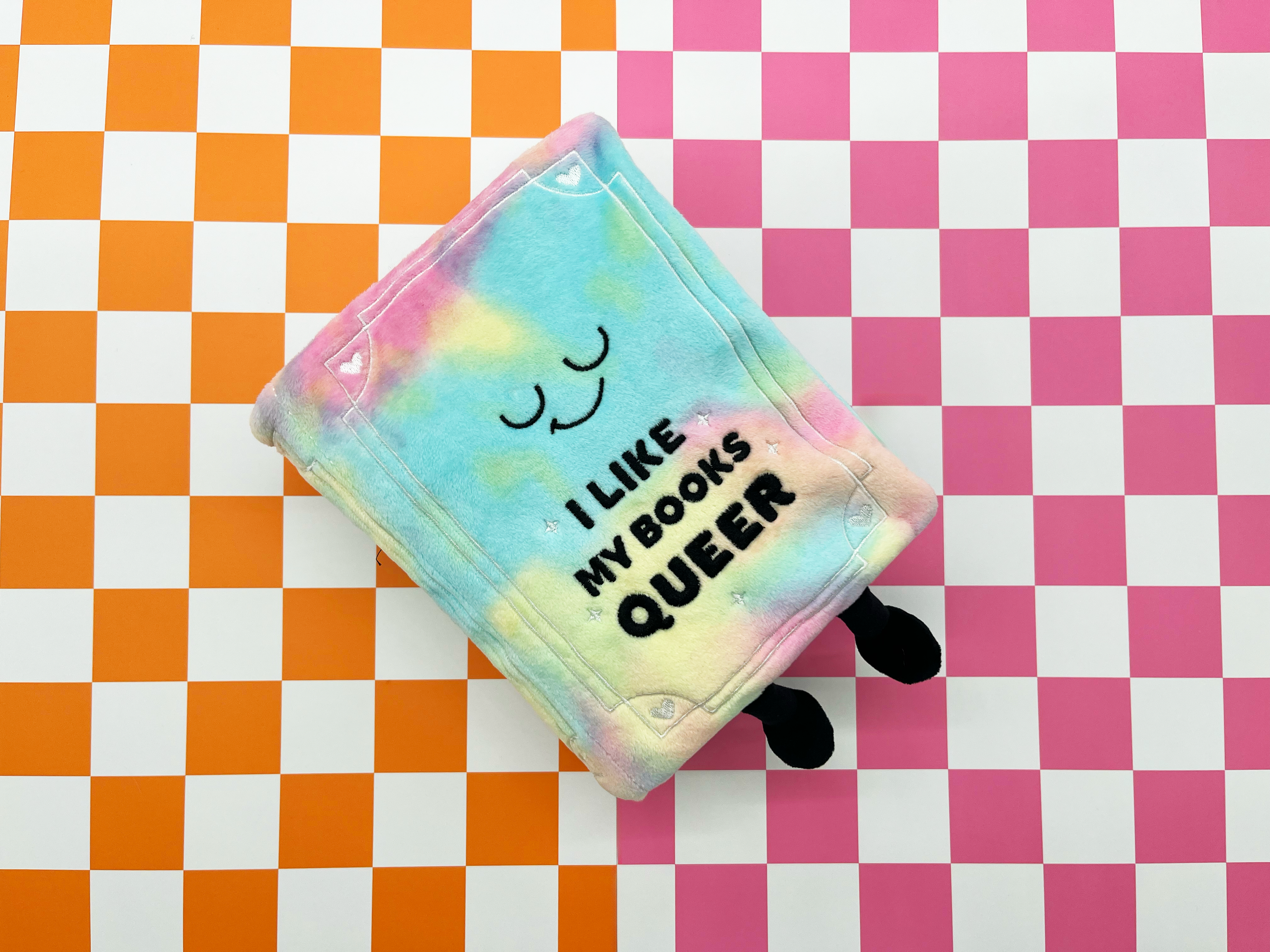 Book plush with text 'I LIKE MY BOOKS QUEER' on a checkered background
