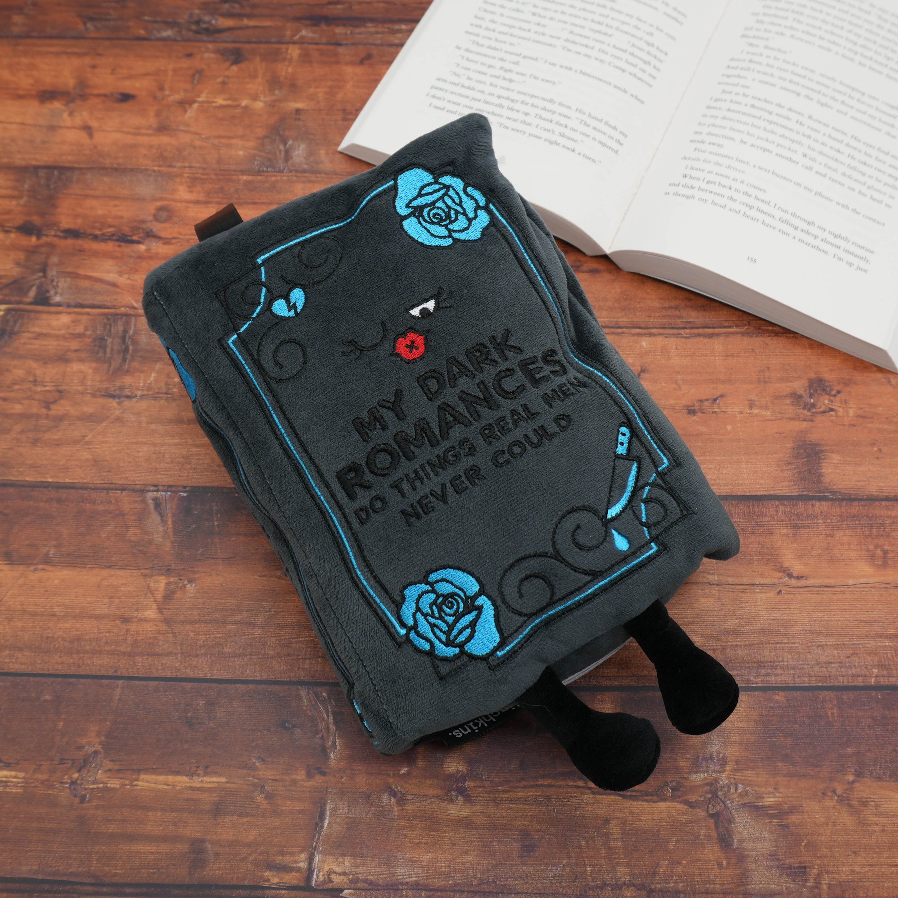 Book-shaped dark romance plush with text and roses on a wooden surface next to an open book.