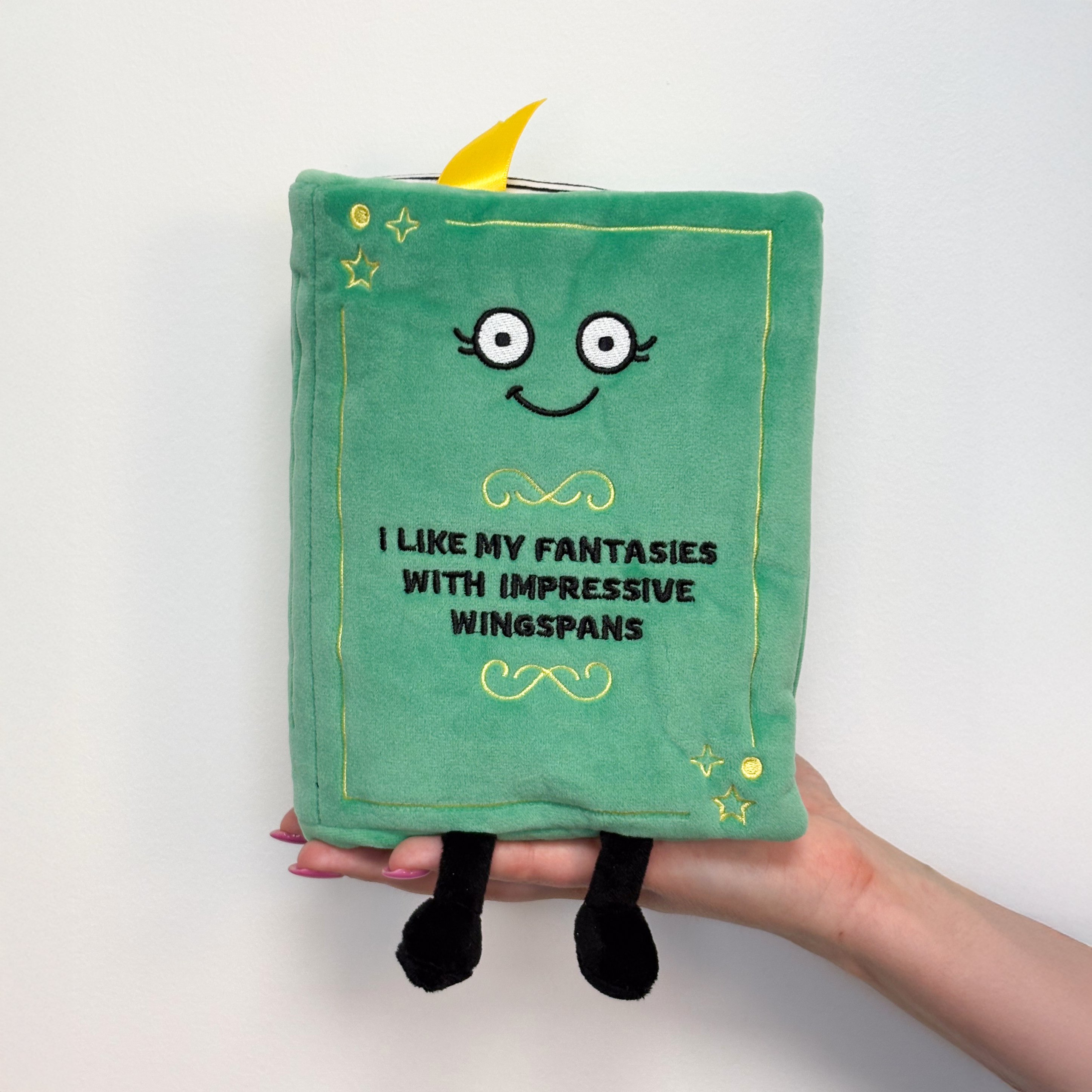 A hand holding a plush designed to look like a green book with a smiling face and 'I like my fantasies with impressive wingspans' written on the cover.