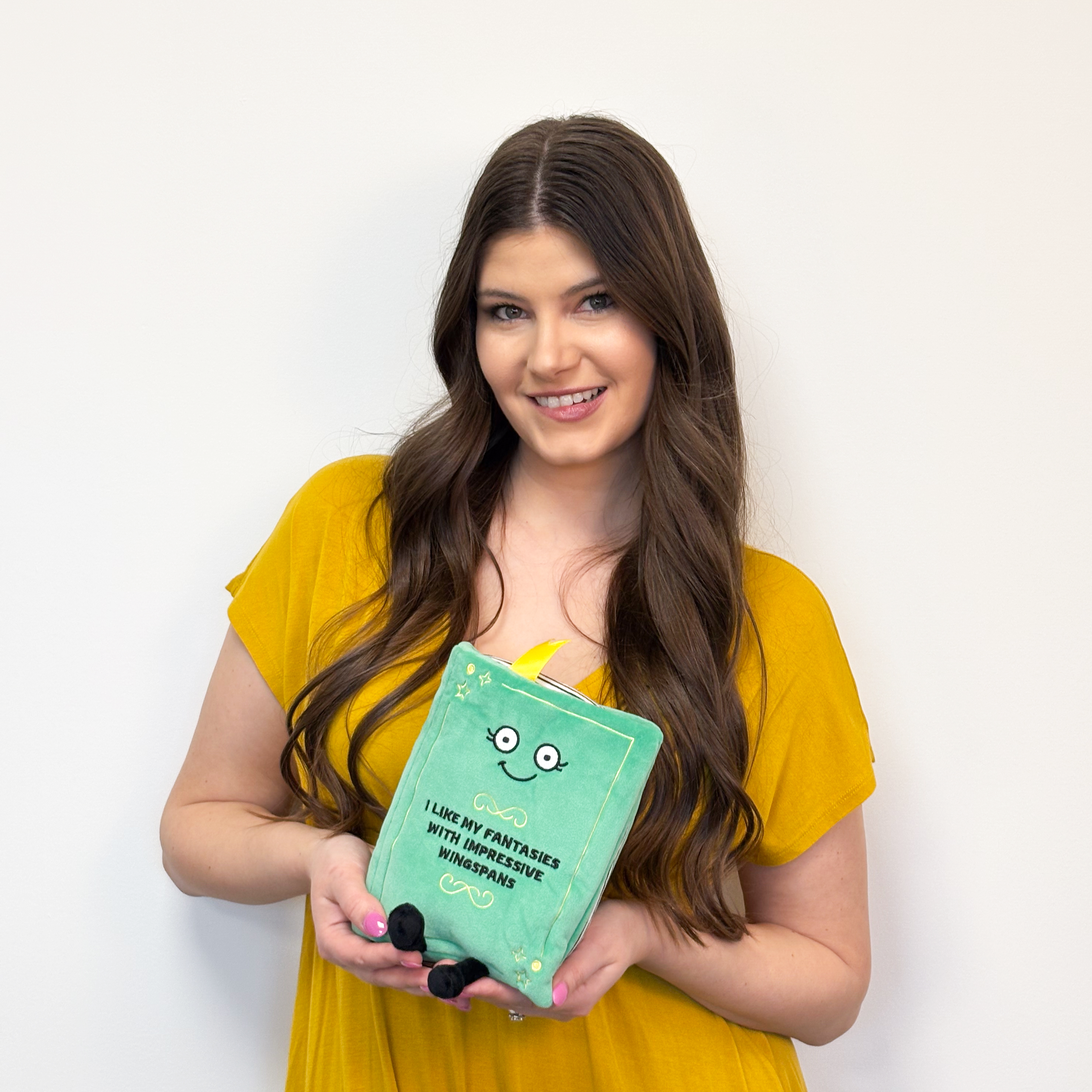 Woman holding a green book plush with a smiling face and 'I like my fantasies with impressive wingspans' written on the cover.