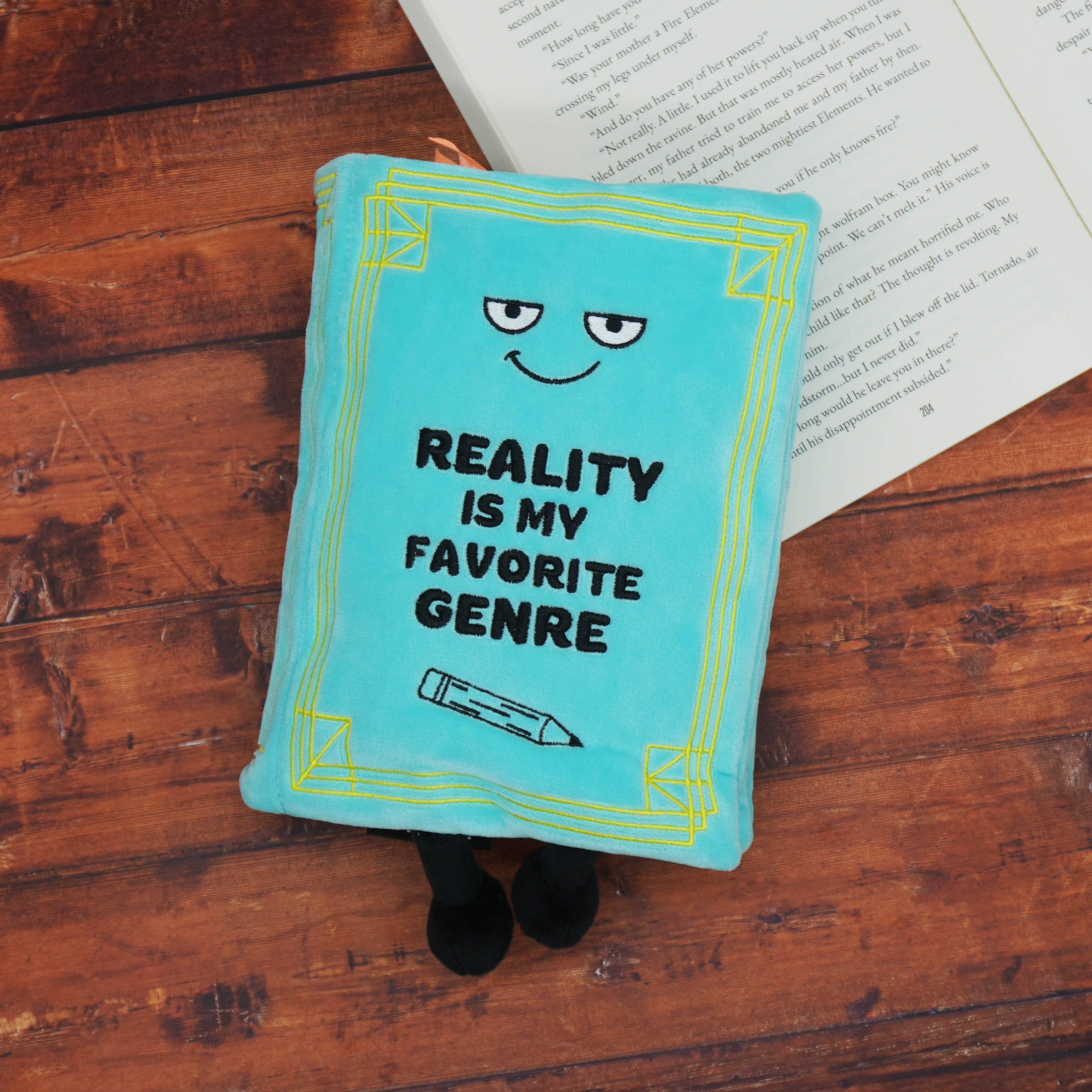 Blue book plush with 'Reality is my favorite genre' text on a wooden surface with an open book.
