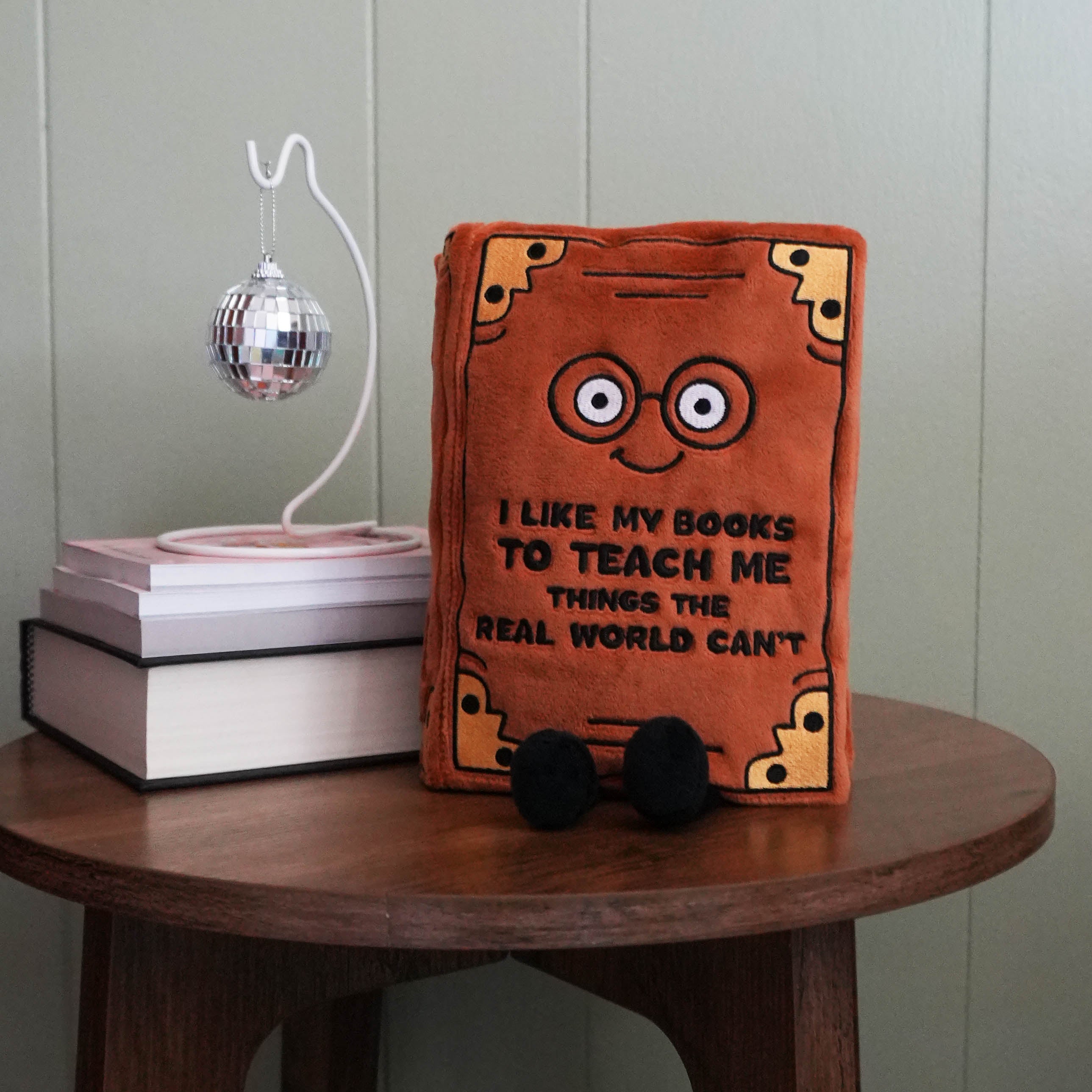 Plush book-shaped toy on a wooden table next to books and a disco ball.