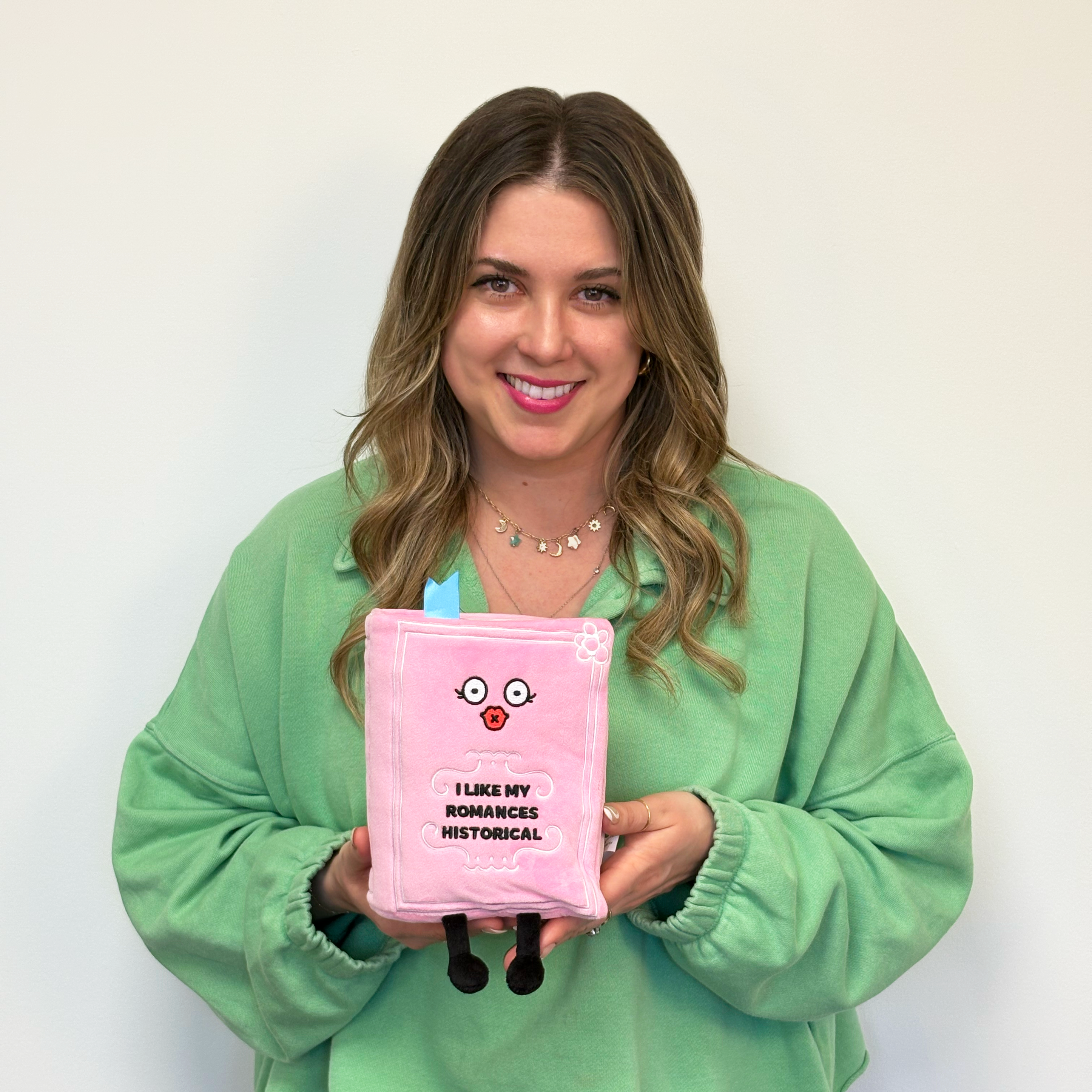 A woman holding a pink plush designed to look like a book with a face, dangly legs, and text that reads, 'I like my romances historical'.