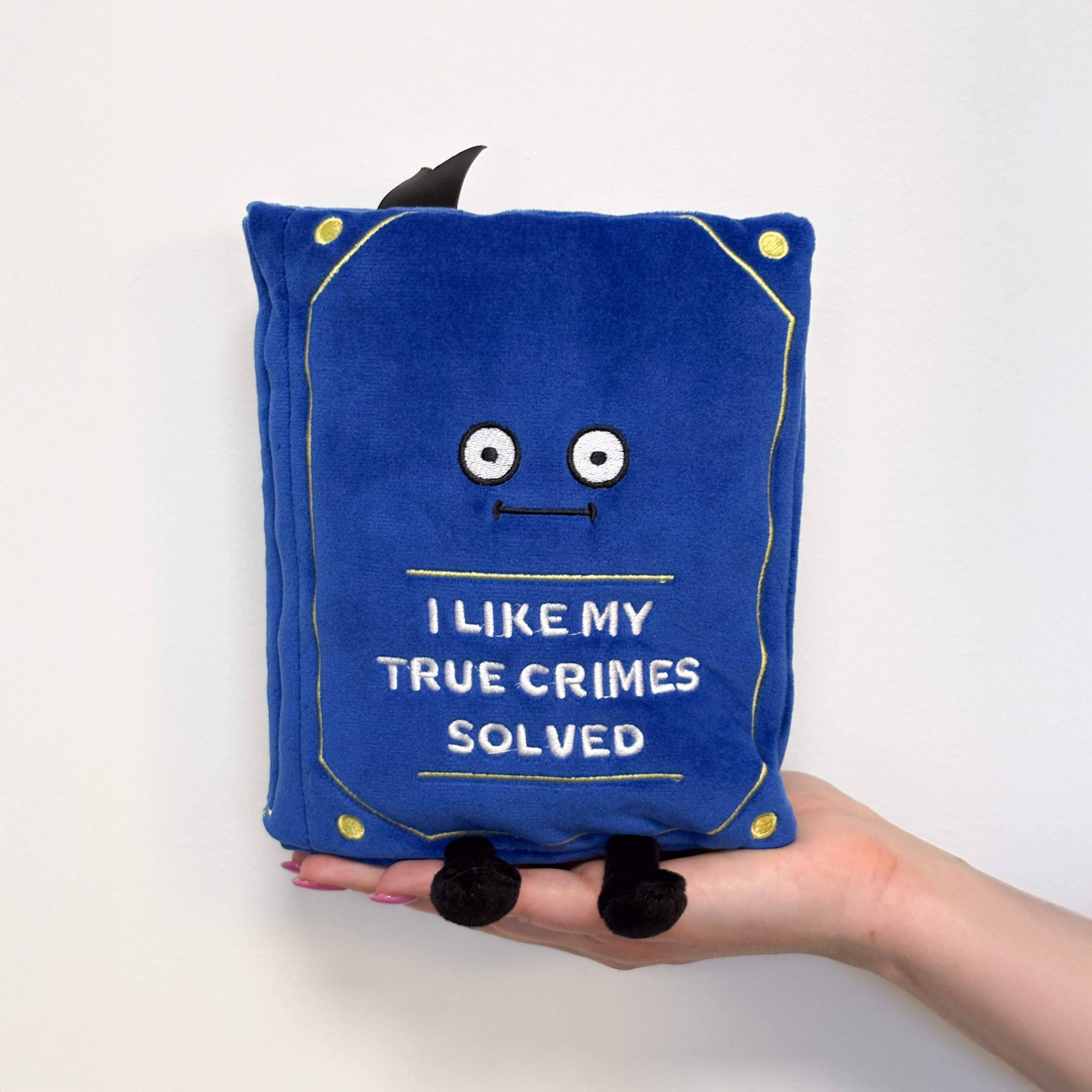 A hand holding a book plush with the title 'I LIKE MY TRUE CRIMES SOLVED' on the cover.