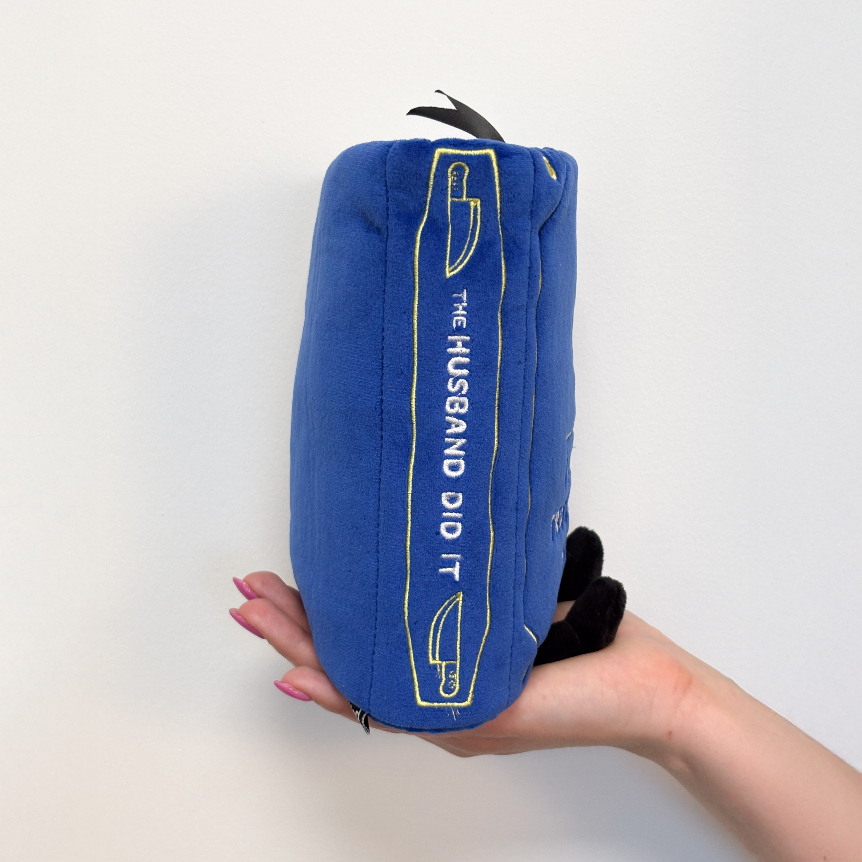 A hand holding a book plush with a spine that reads "The husband did it."