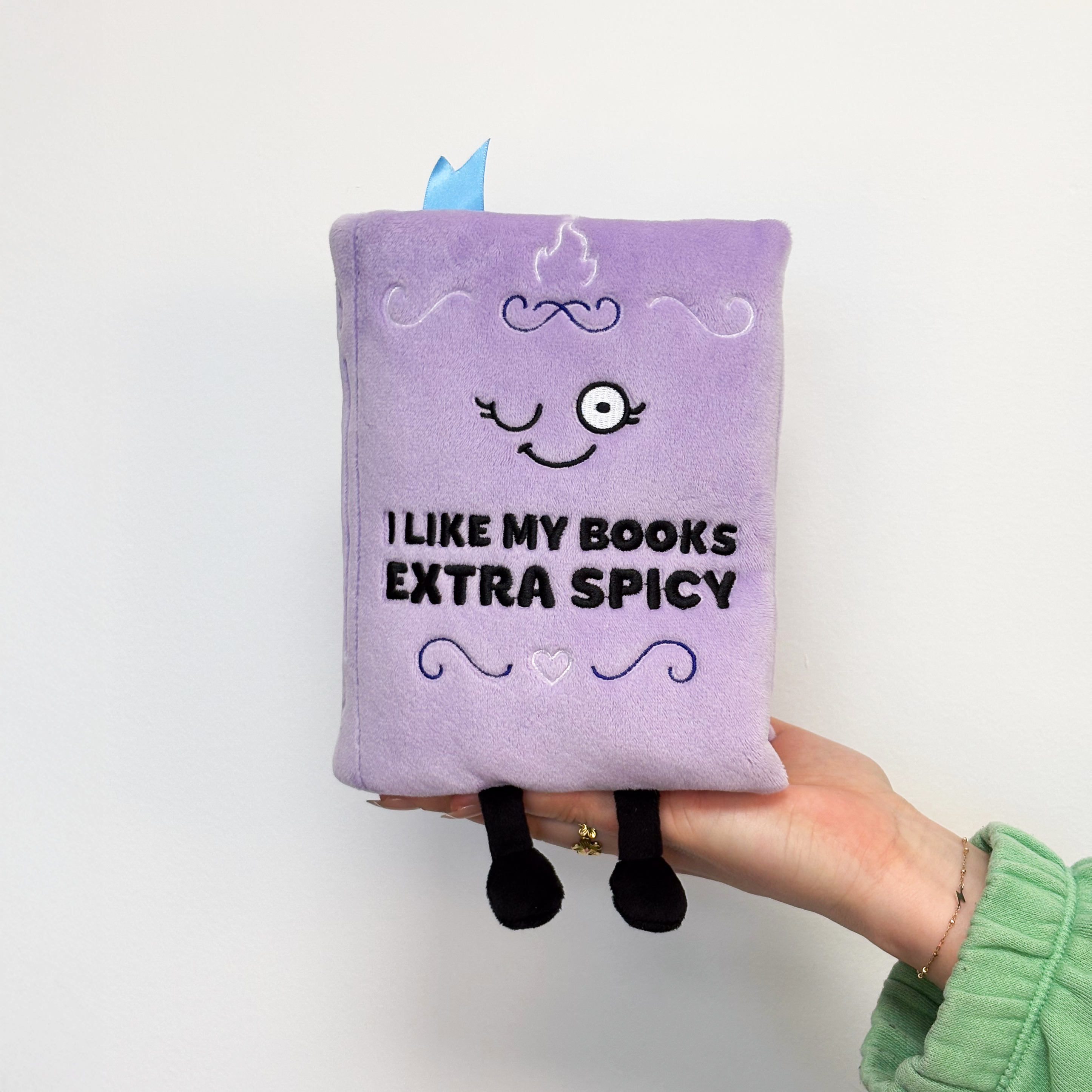A hand holding a plush book with the phrase 'I LIKE MY BOOKS EXTRA SPICY' written on the cover. 