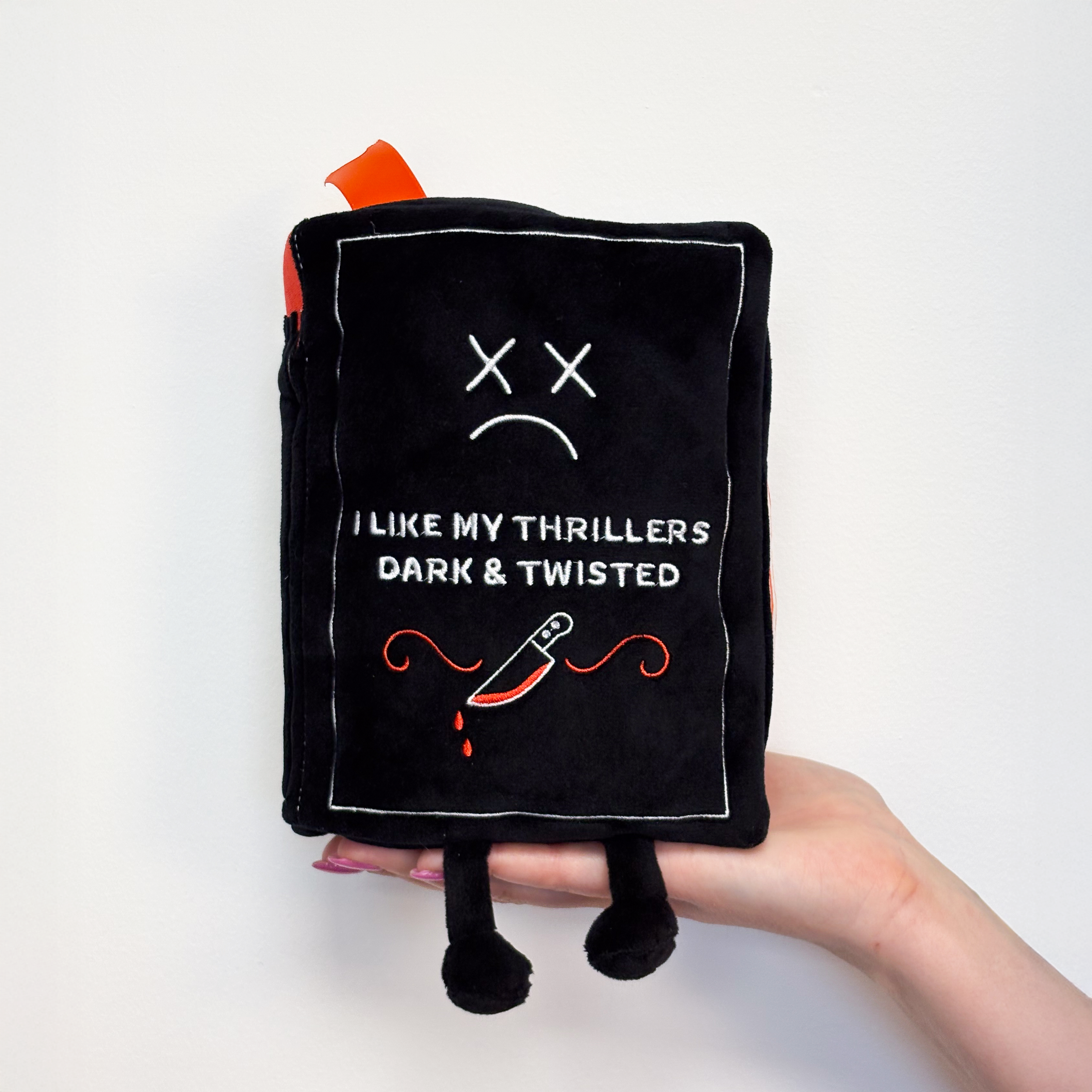A hand holding a plush designed to look like a book with the description 'I Like My Thrillers Dark & Twisted' on the cover.