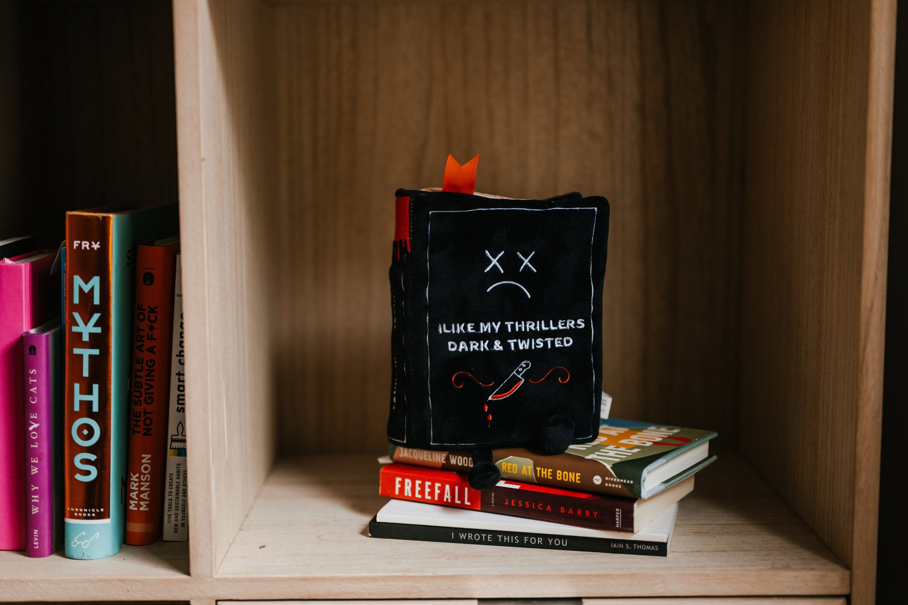 A plush designed to look like a book with the description 'I Like My Thrillers Dark & Twisted' on the cover on a bookshelf. 