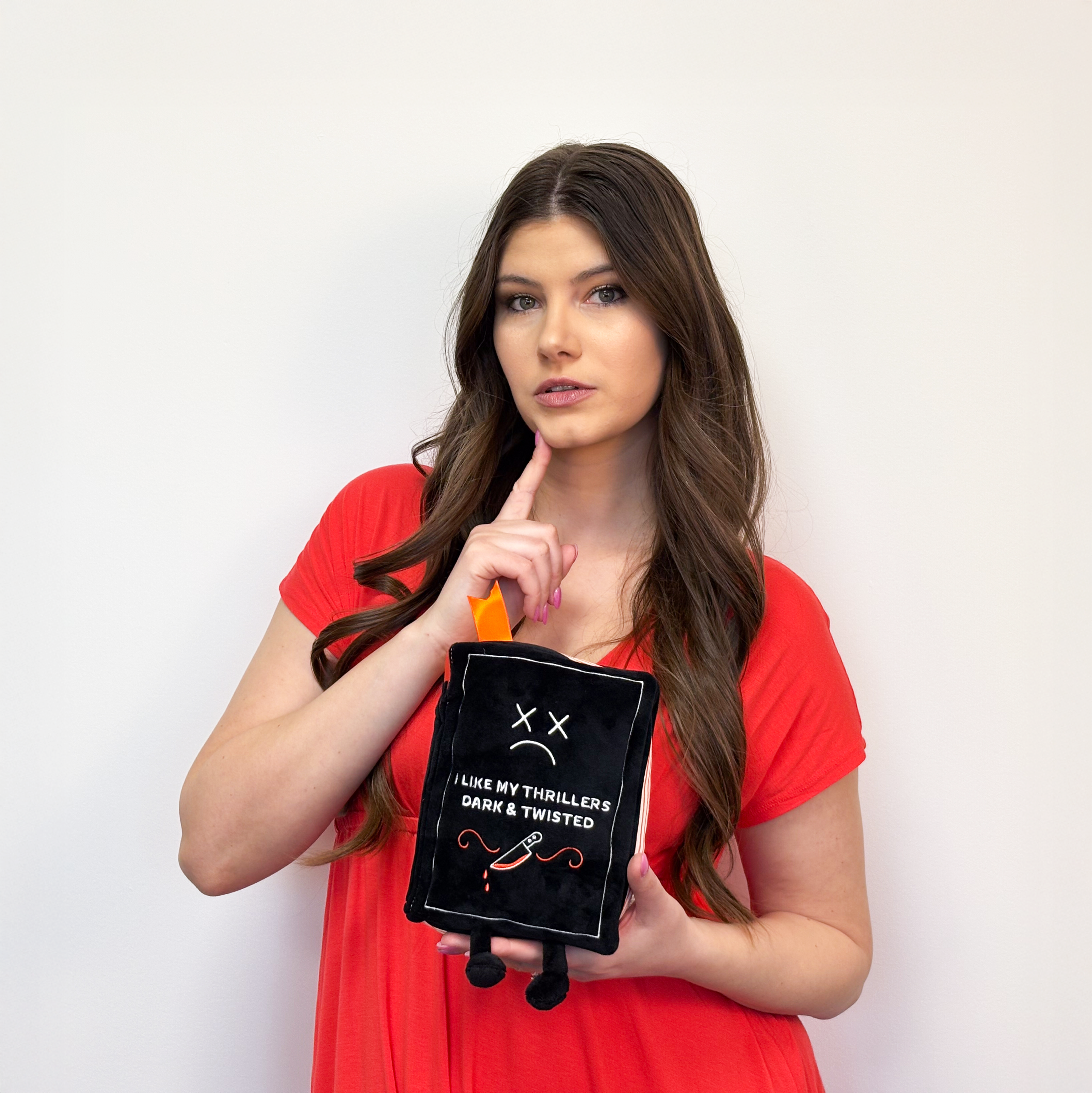 A woman holding a plush designed to look like a book with the description 'I Like My Thrillers Dark & Twisted' on the cover.