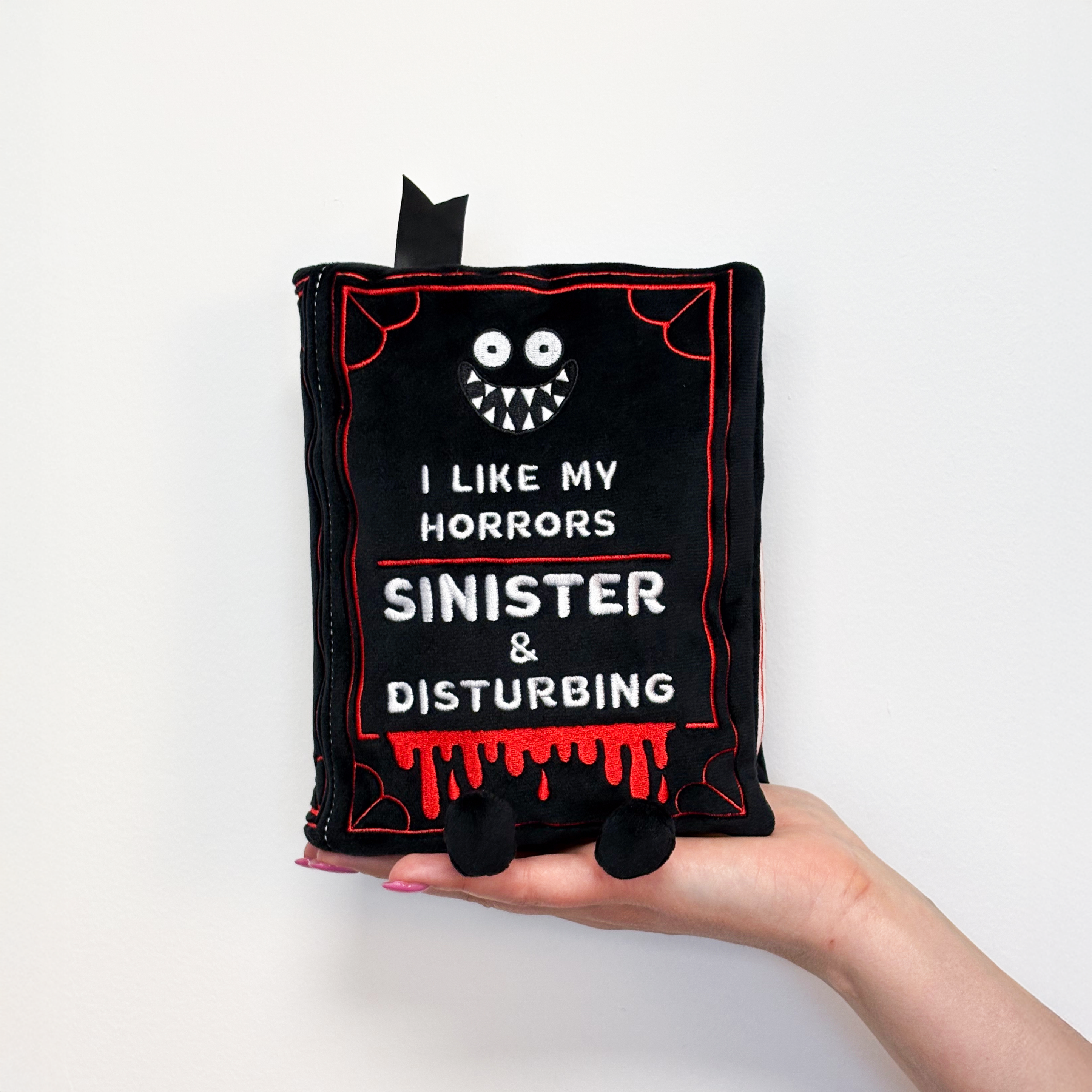 Hand holding a plush book featuring the text 'I LIKE MY HORRORS SINISTER & DISTURBING' on the front. 