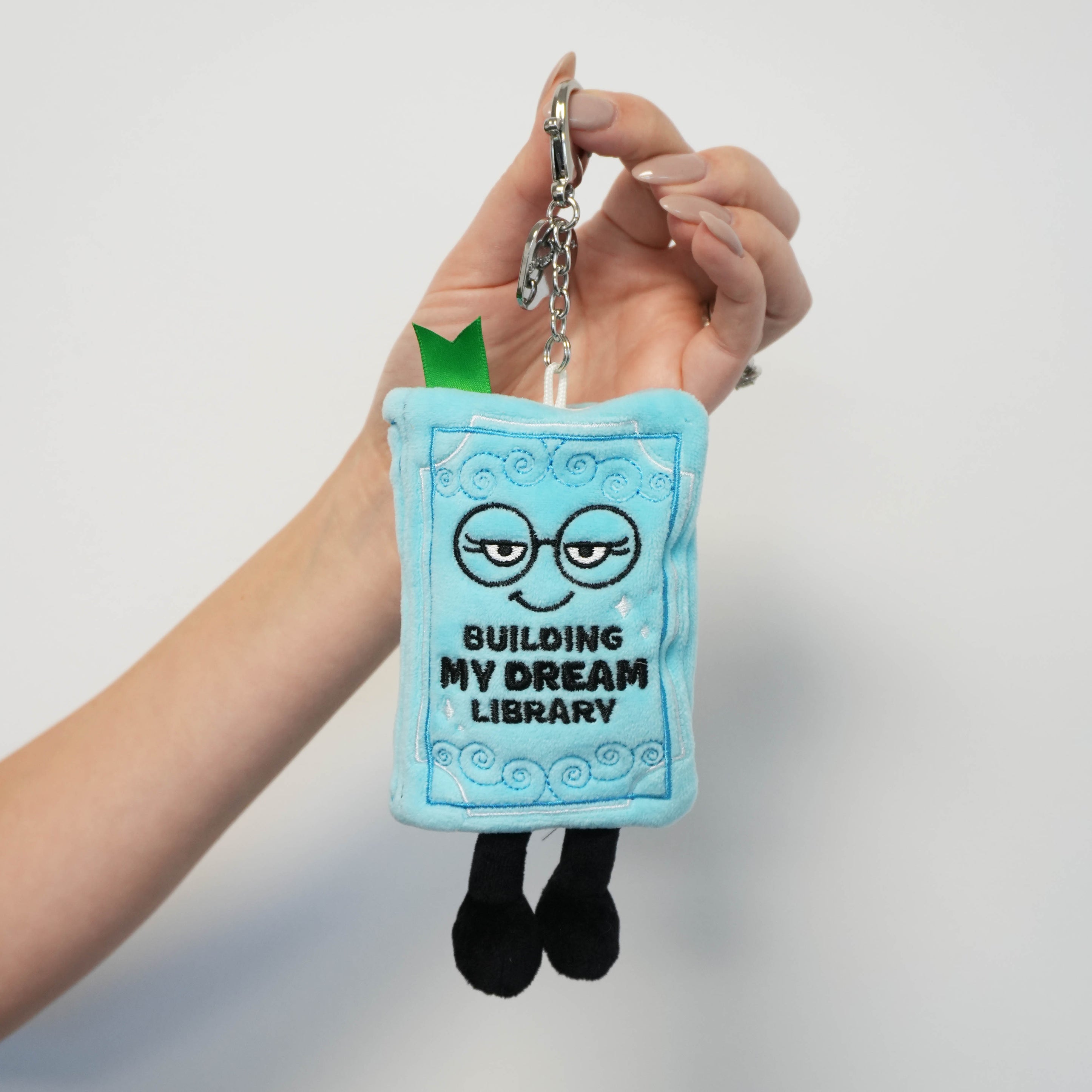 Plush keychain with 'Building My Dream Library' text held by a hand against a light gray background