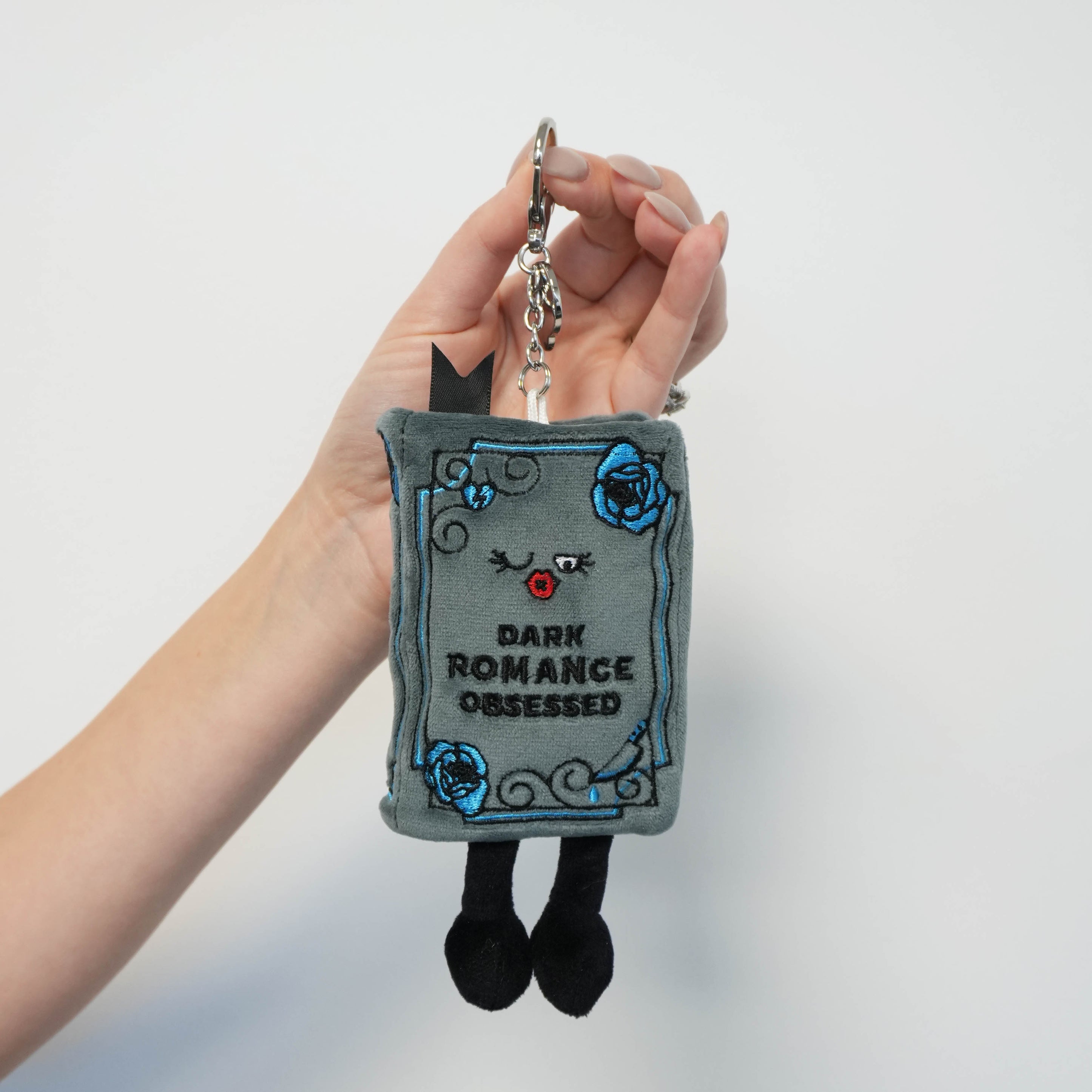 Plush keychain shaped like a book titled 'Dark Romance Obsessed' held by a hand against a light gray background.