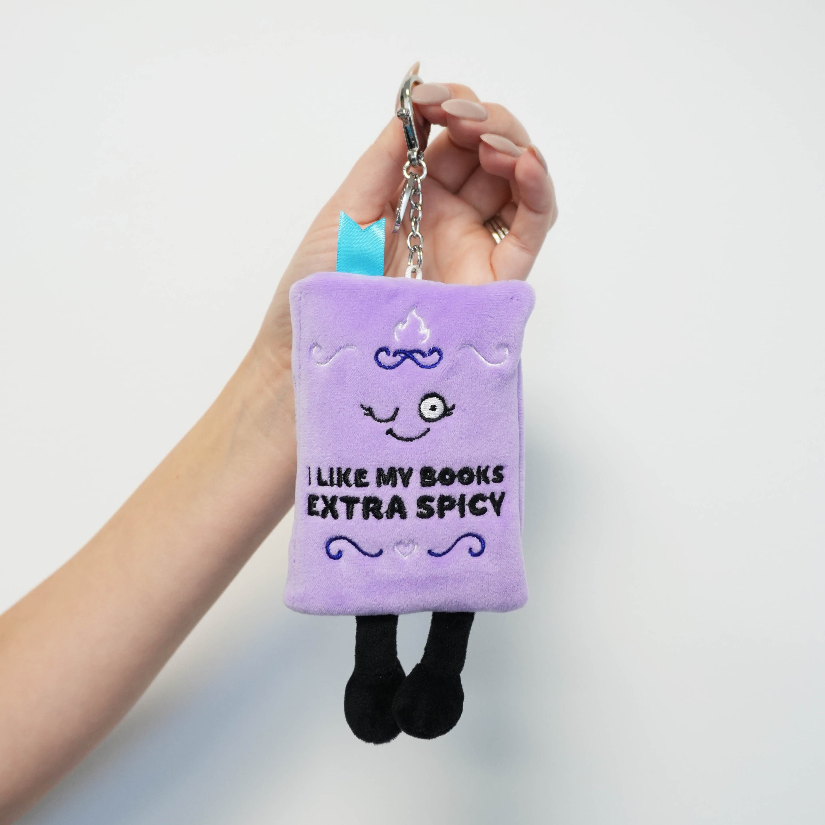 Plush book bag charm with 'I like my books extra spicy' text held by a hand against a light gray background