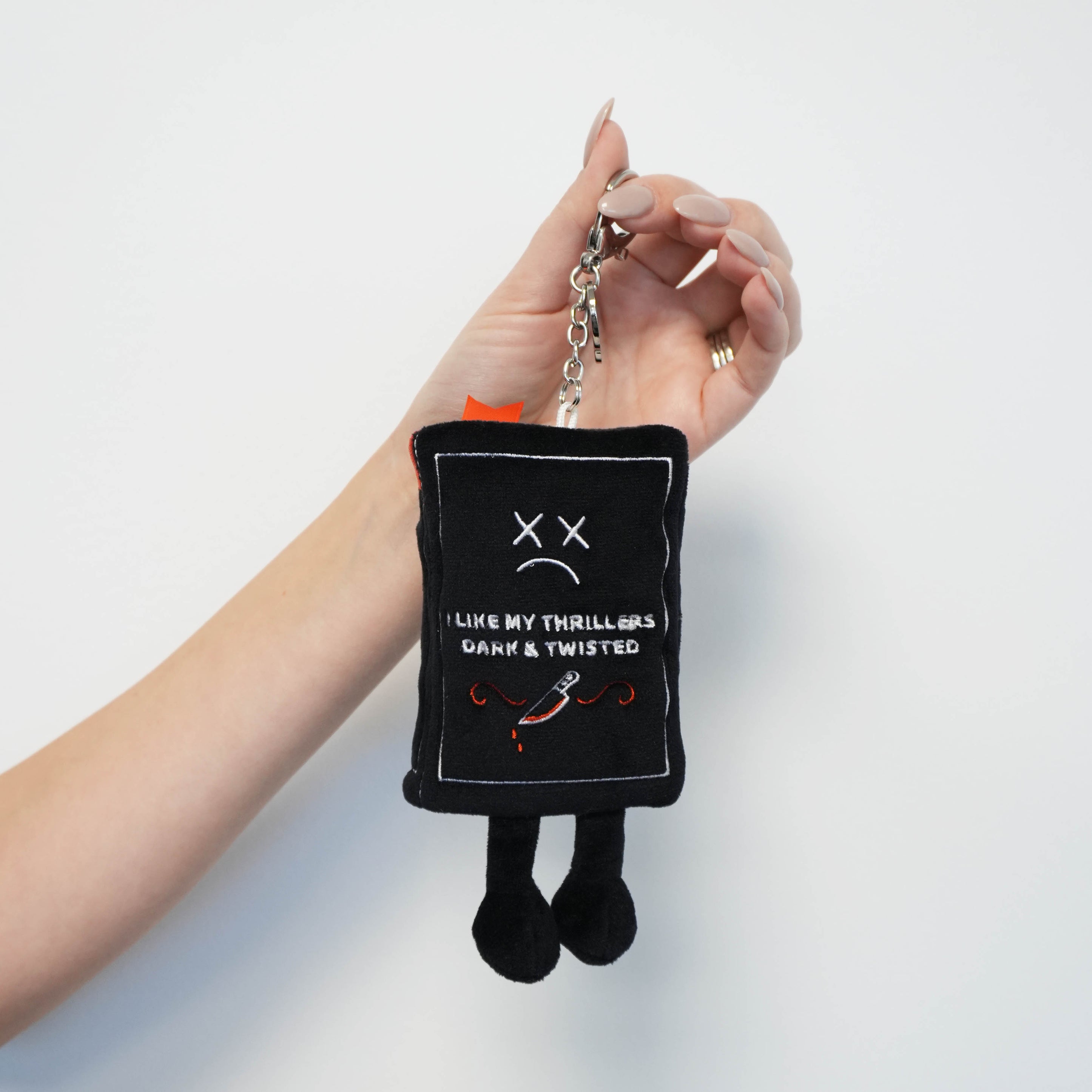 Black book plush bag chain with the phrase 'I like my thrillers dark & twisted' held by a hand against a white background