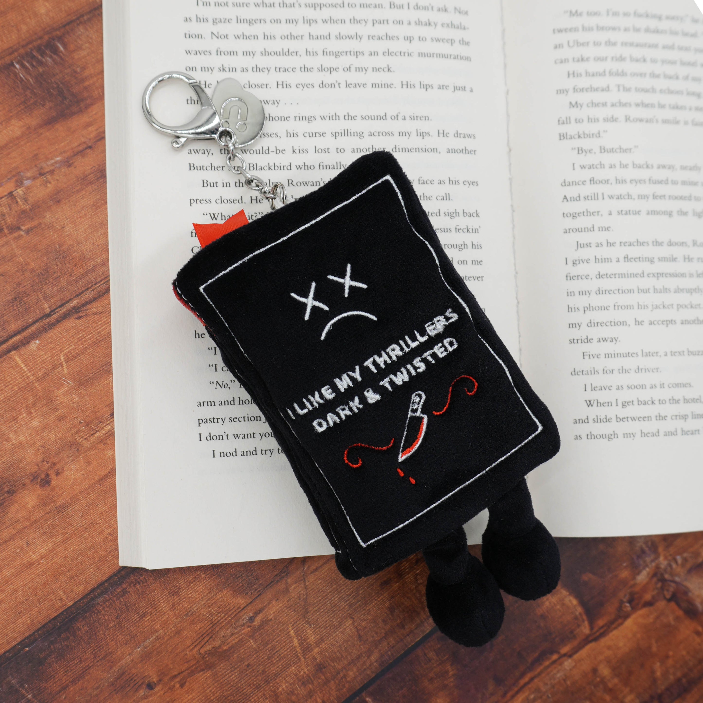 Black plush book keychain with 'I like my thrillers dark & twisted' written on it placed on a book