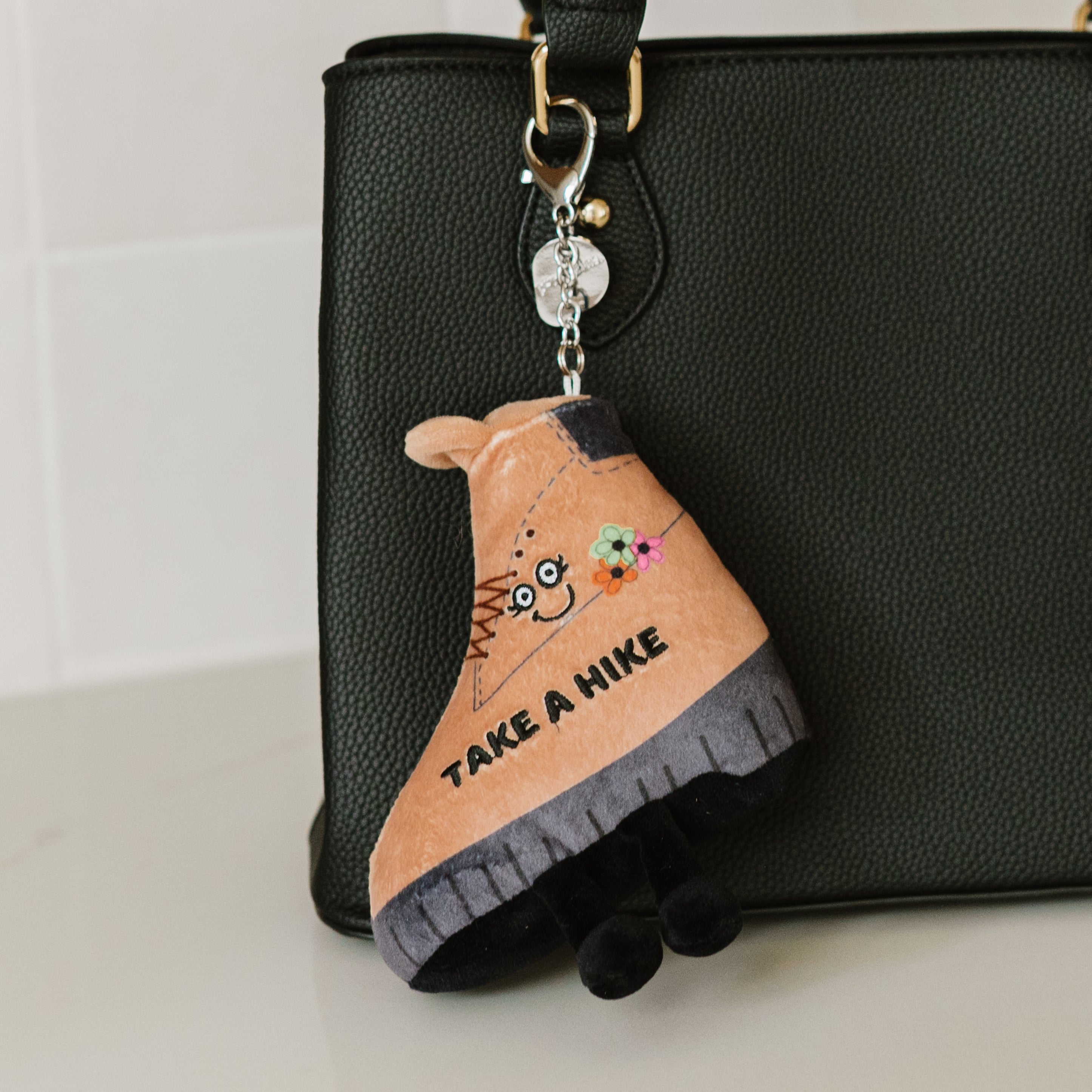A plush bag charm shaped like a brown hiking boot with a smiling face and the phrase 'TAKE A HIKE' on it hanging off a purse.