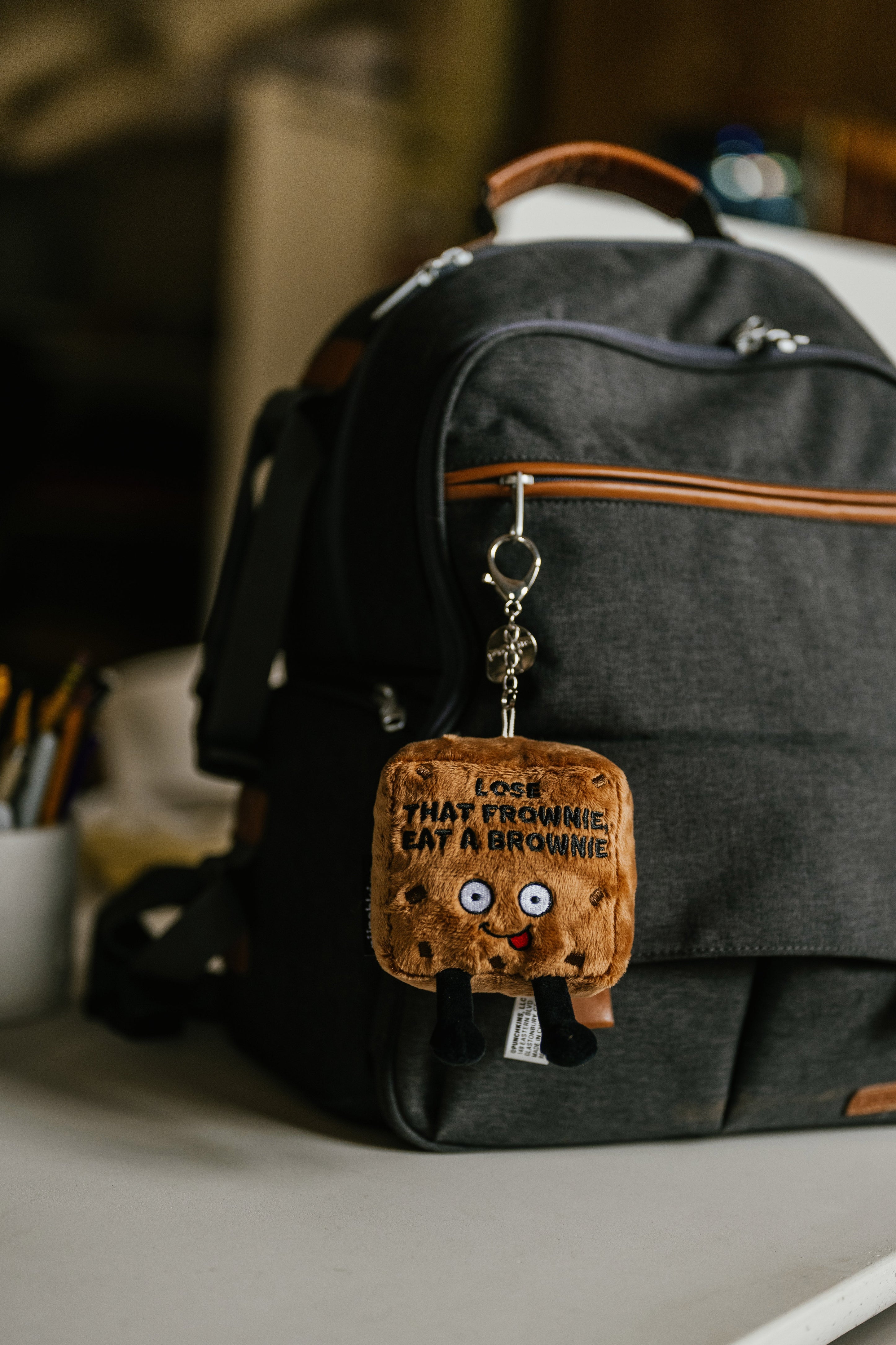 A plush brownie-shaped bag charm with the text that reads 'LOSE THAT FROWNIE, EAT A BROWNIE' hanging on a backpack.