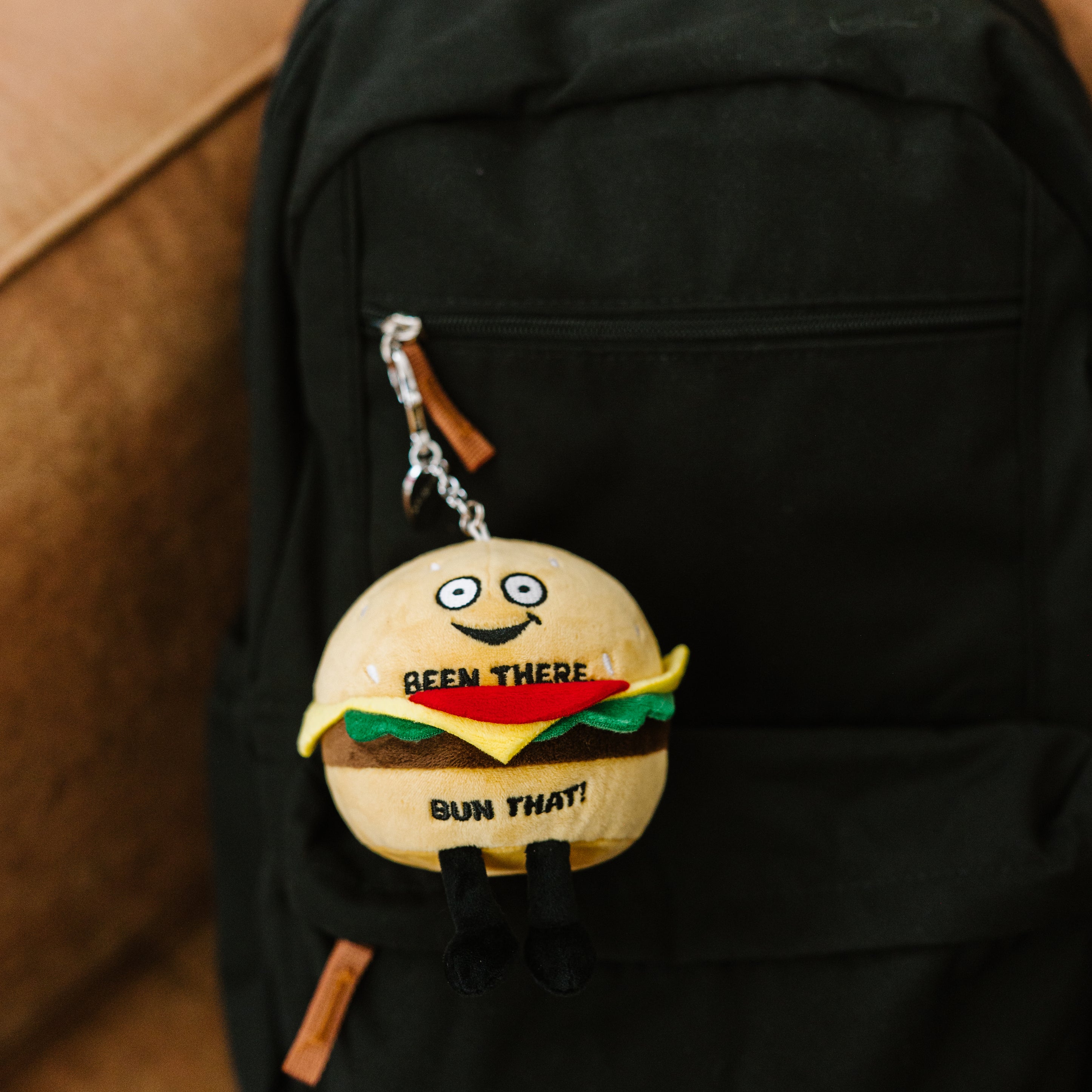 A plush bag charm shaped like a burger with a smiling face and the text 'BEEN THERE, BUN THAT!' written on it hanging off a backpack.