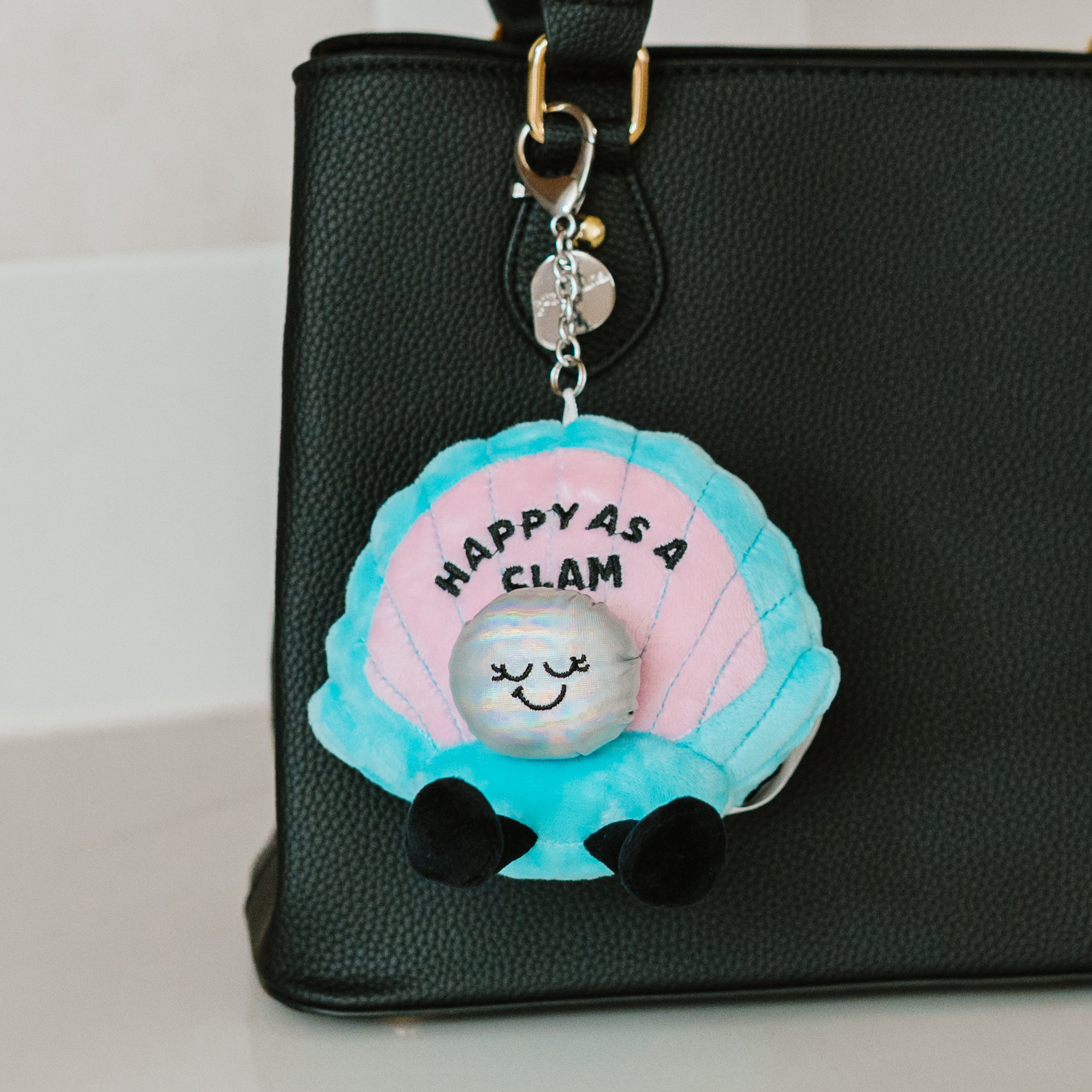 A plush charm designed to look like a clam, featuring a pearl accent and the text 'HAPPY AS A CLAM' on its shell hanging off a purse.