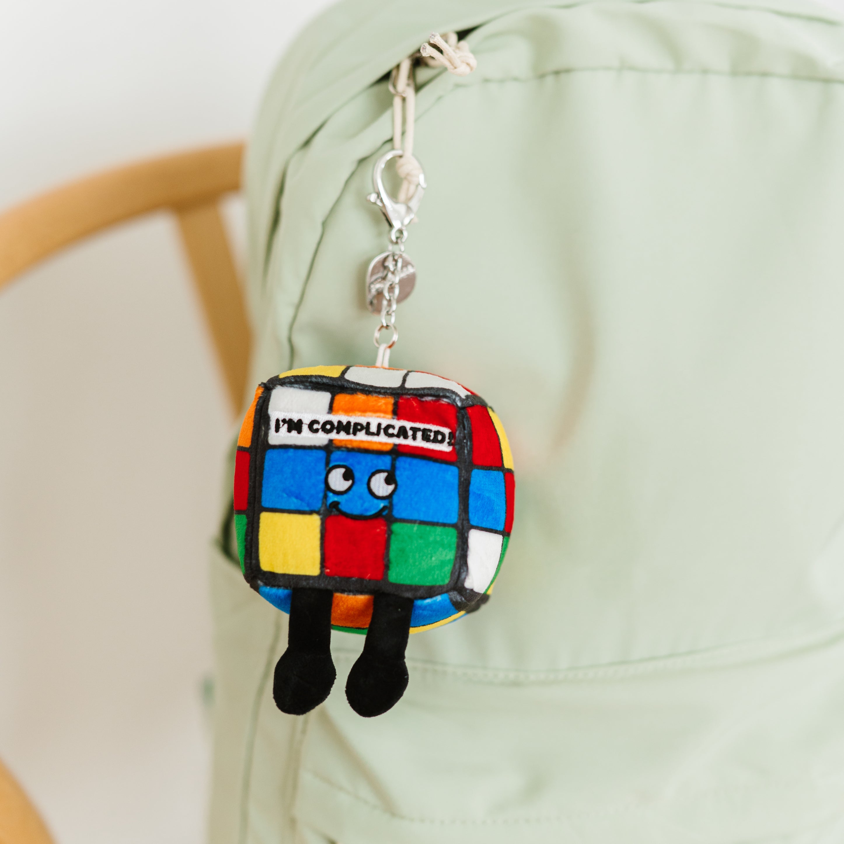 A colorful cube-shaped plush bag charm with character features and the text 'I'M COMPLICATED!' on the front hanging off a backpack. 
