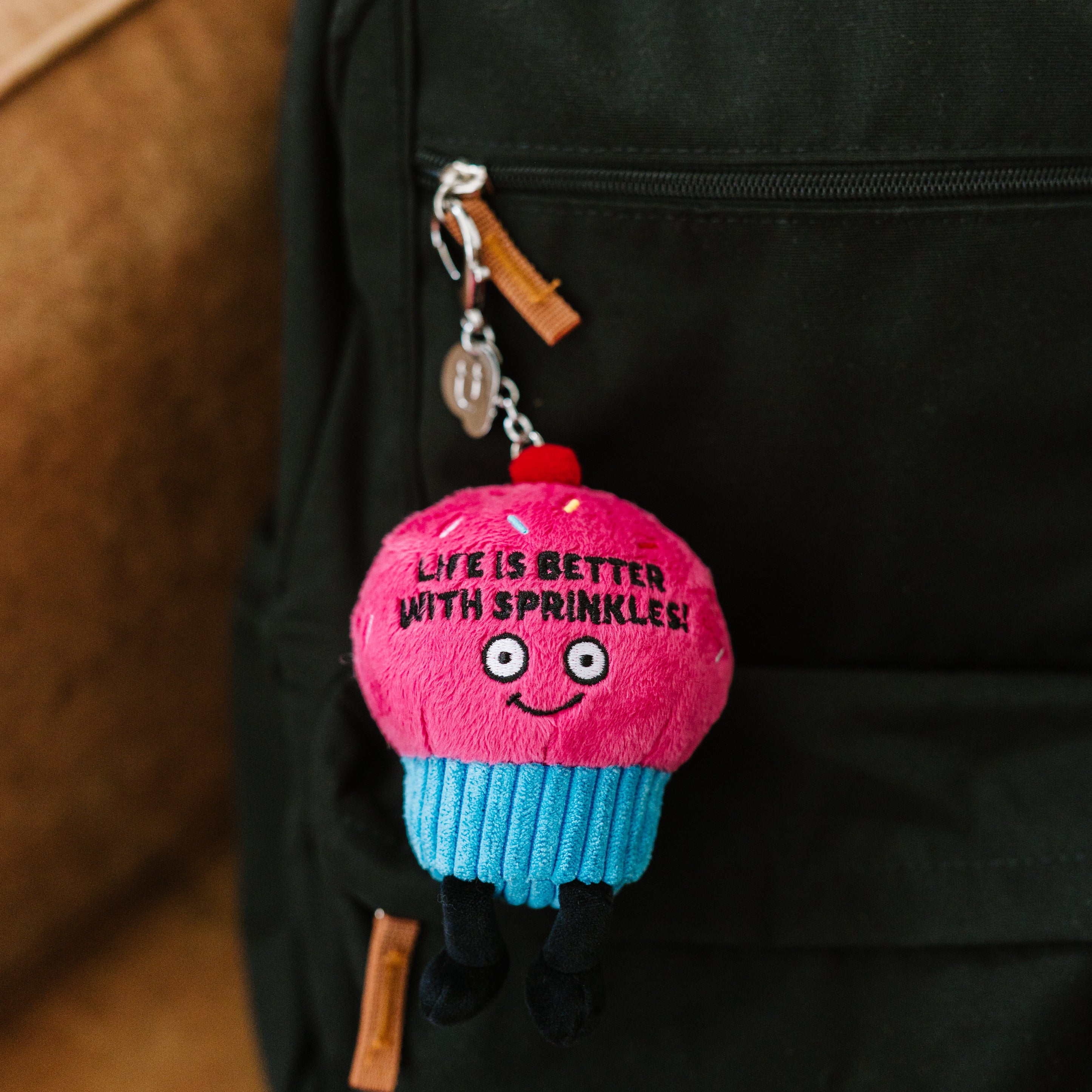A plush charm designed to look like a cupcake with the text 'Life is better with sprinkles' written on it hanging off a backpack.