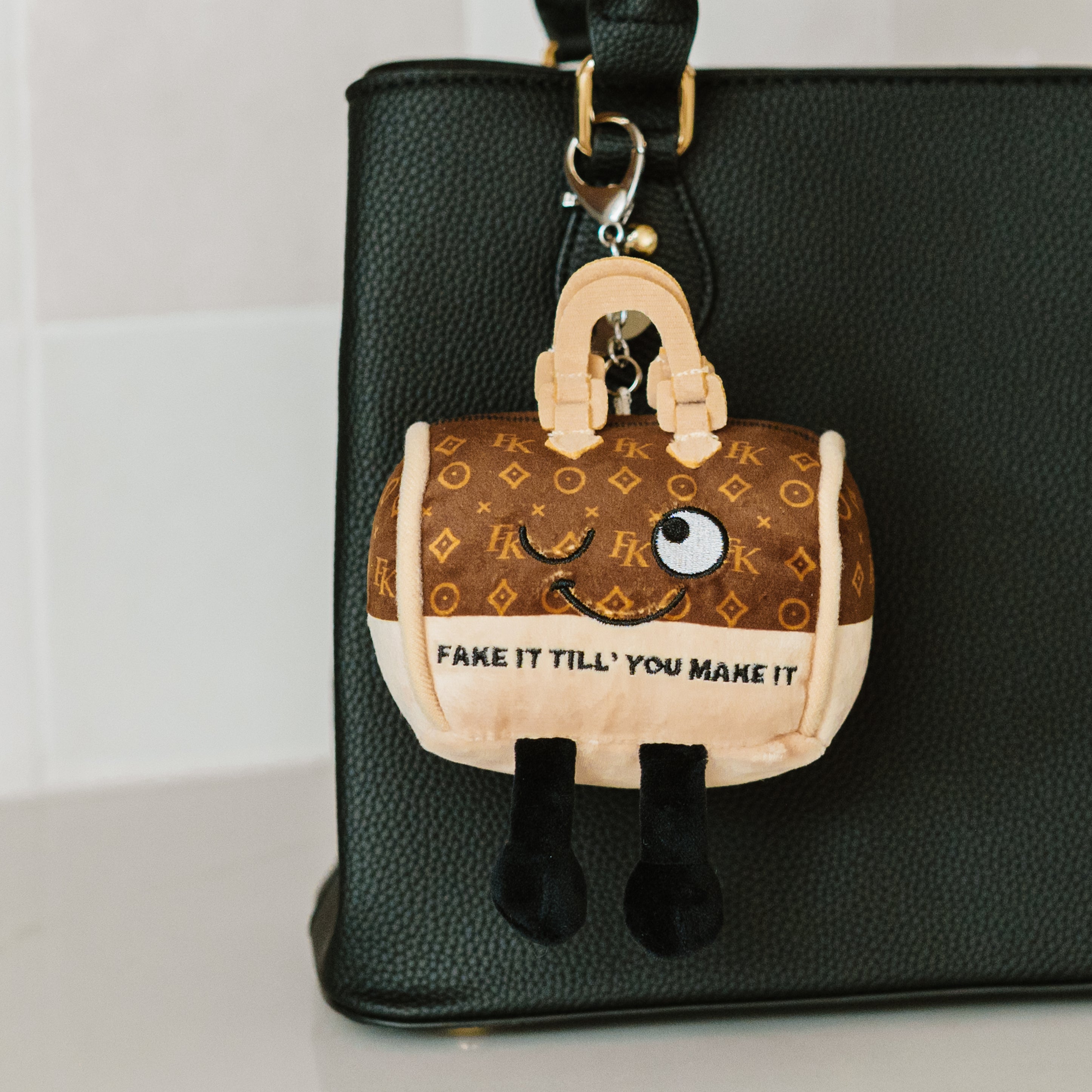 A plush bag charm of a purse featuring a whimsical face and text that reads, "Fake it till you make it" hanging off a purse.
