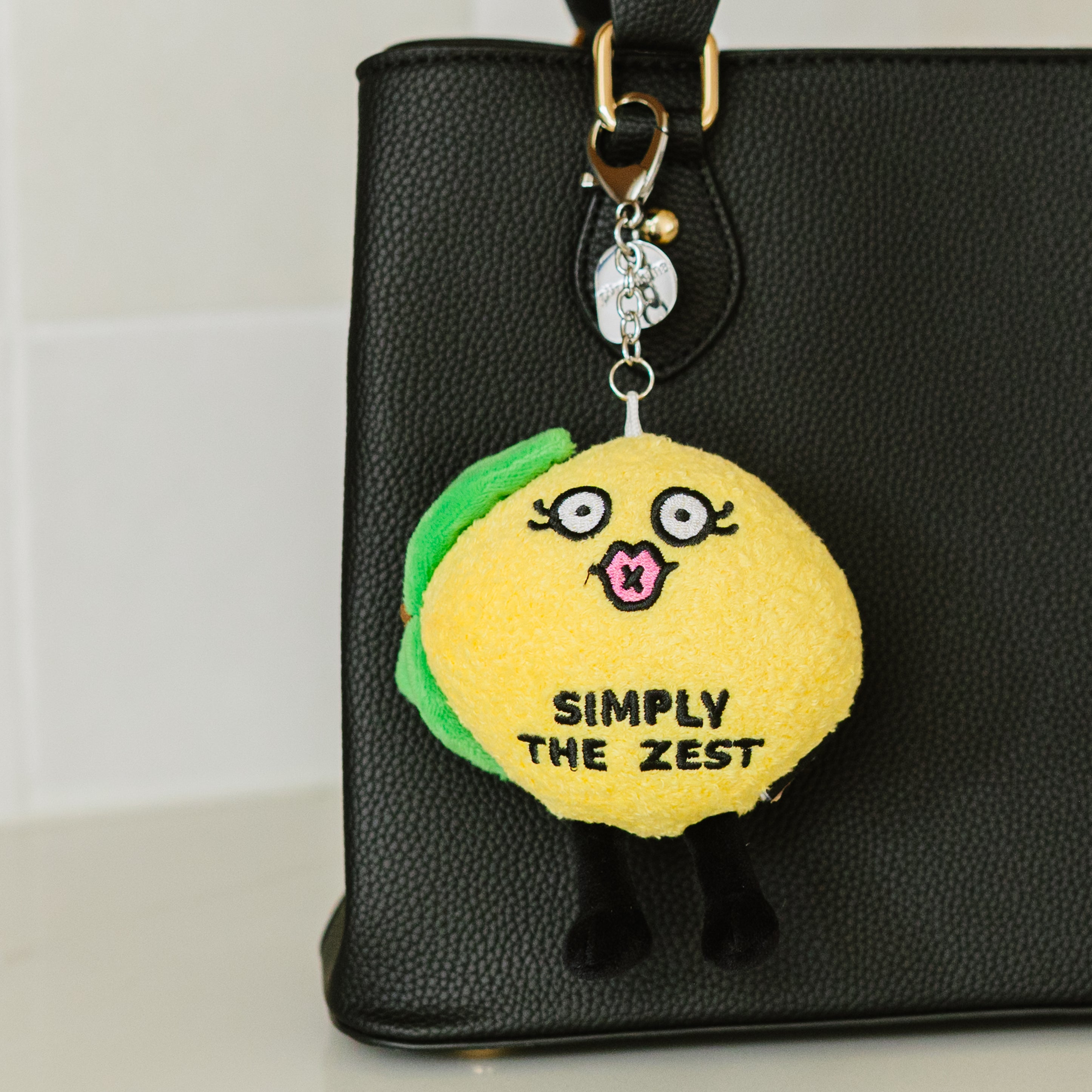 A plush lemon bag charm with a face, legs, and the text 'SIMPLY THE ZEST' printed on it hanging off a purse.