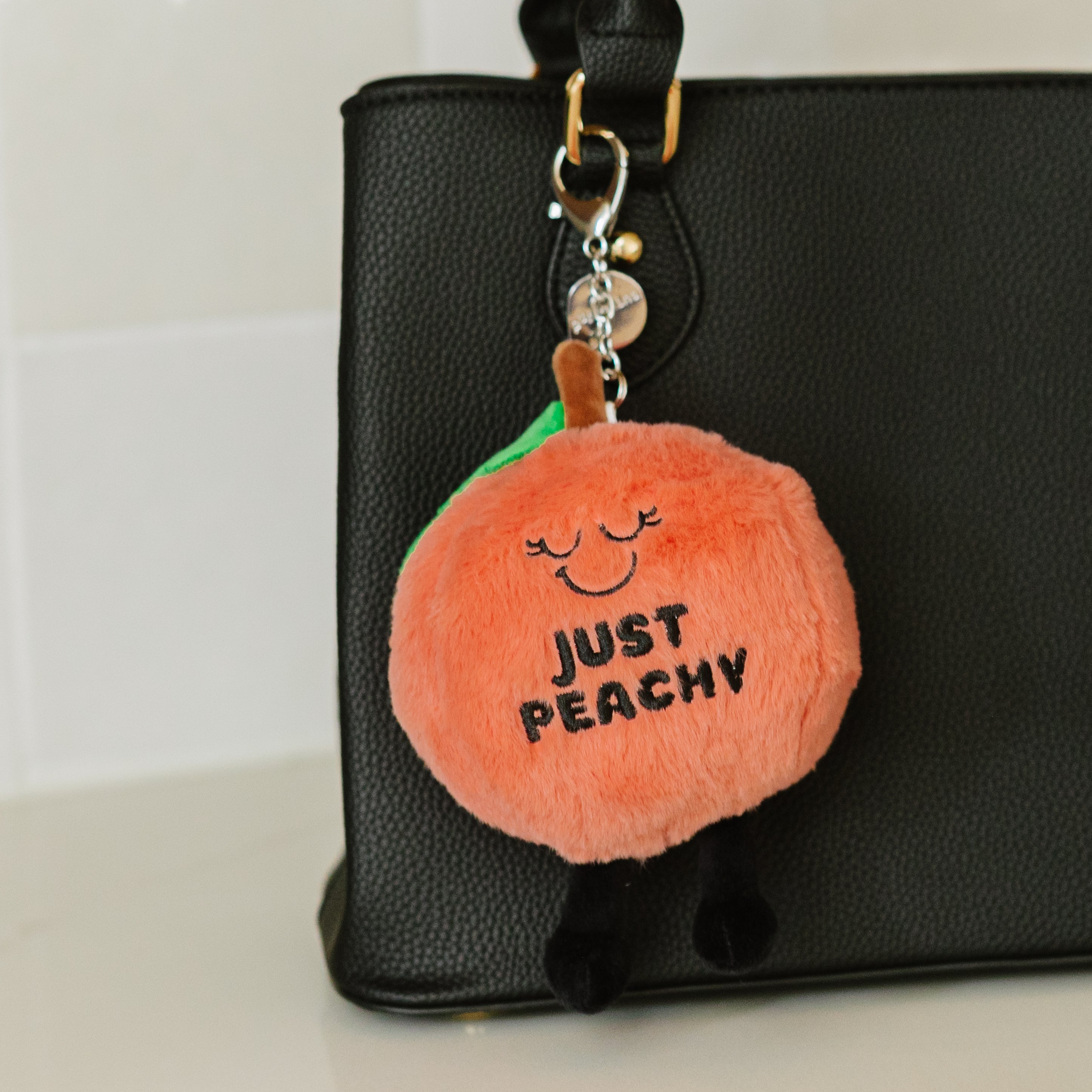 A plush peach bag charm with a smiling face, eyes, dangly legs, and the text 'JUST PEACHY' printed on it hanging off a purse.